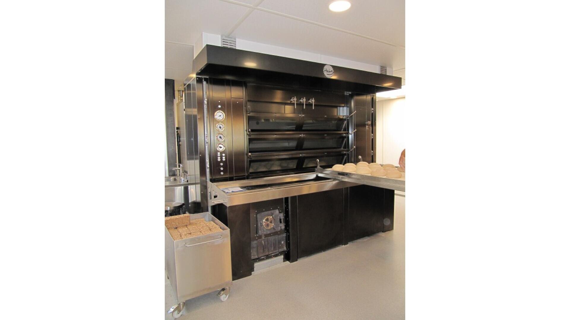 A large, black commercial bread oven with multiple shelves stands in a bakery. A metal table in front holds several unbaked dough loaves, and a cart with a tray of baked bread sits nearby.