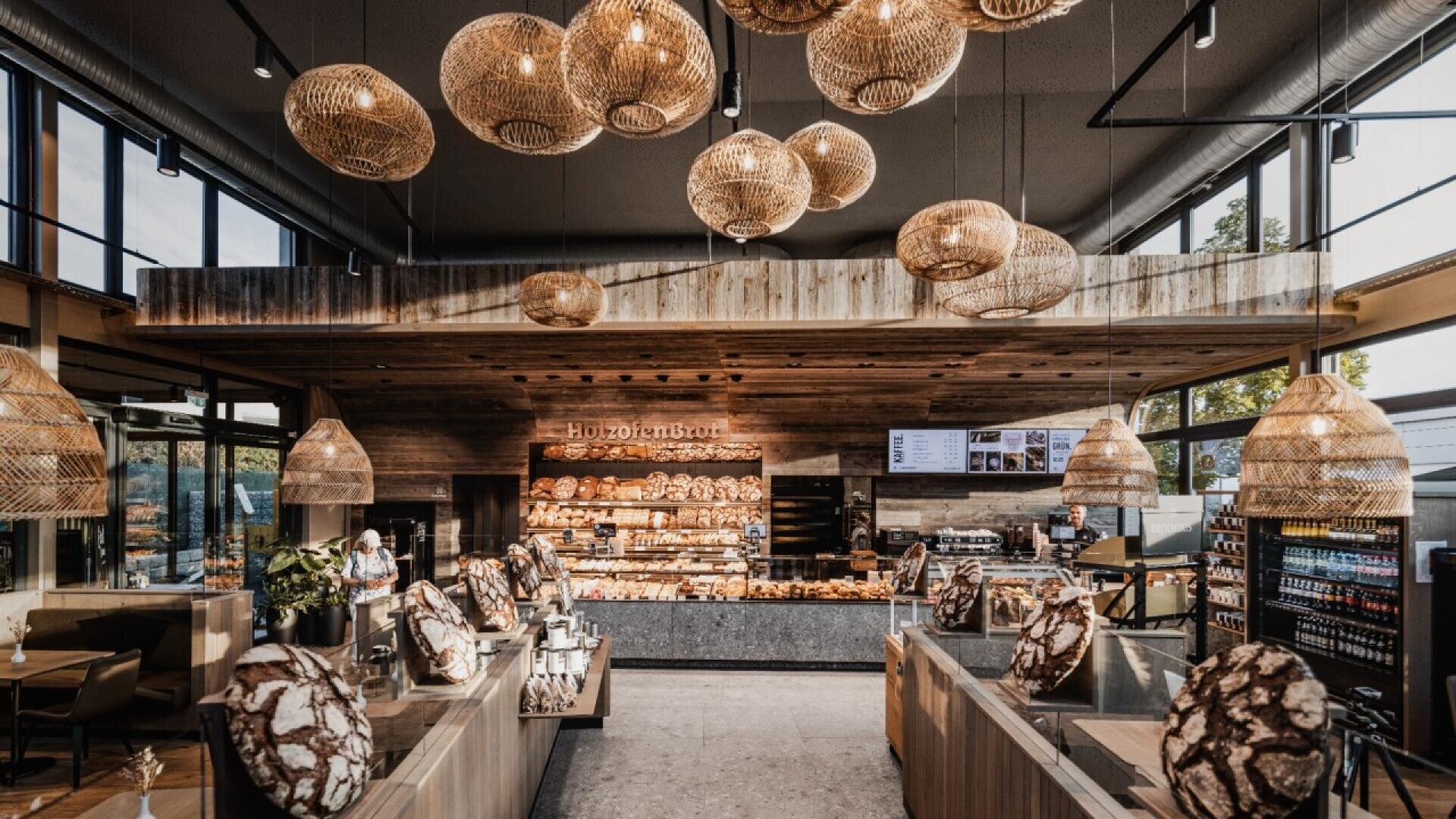 A modern bakery with rustic wooden decor, large wicker pendant lights, display cases filled with bread and pastries, and large windows letting in natural light.