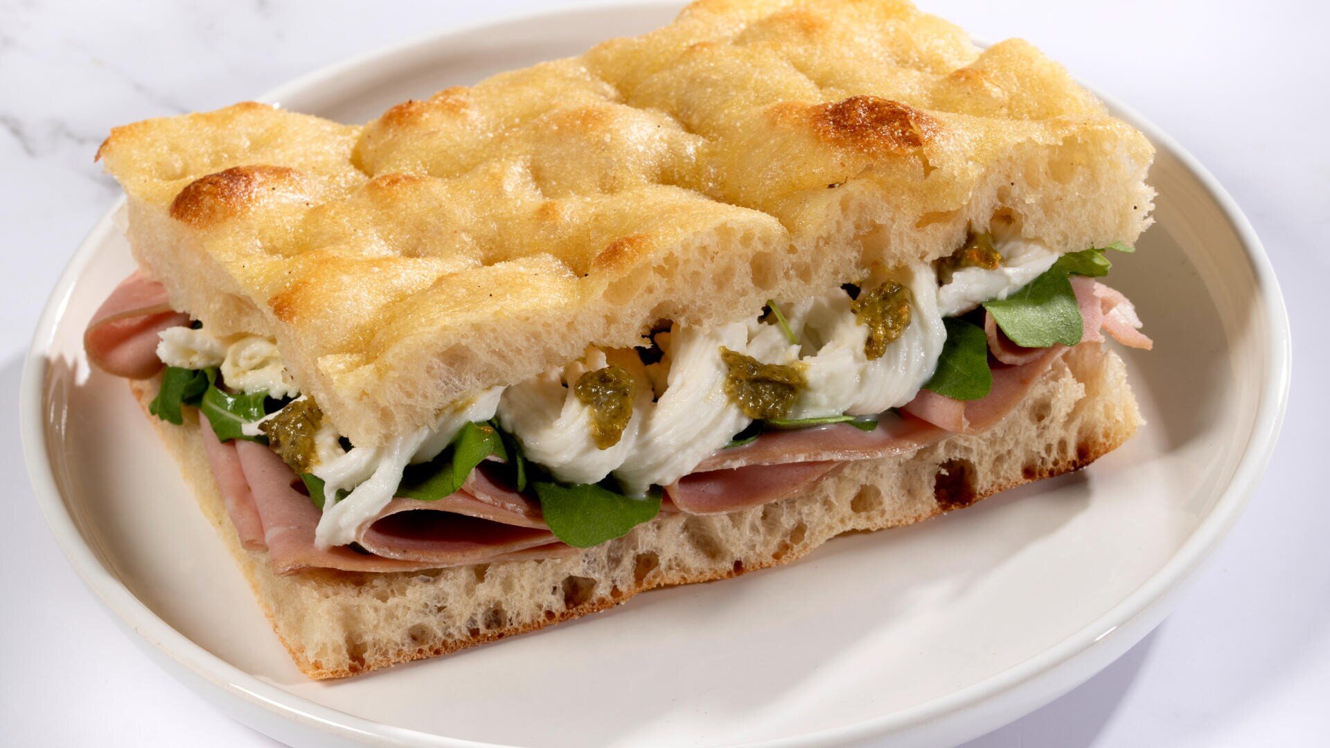A sandwich made with focaccia bread filled with sliced ham, mozzarella cheese, arugula, and a green pesto spread, served on a white plate.