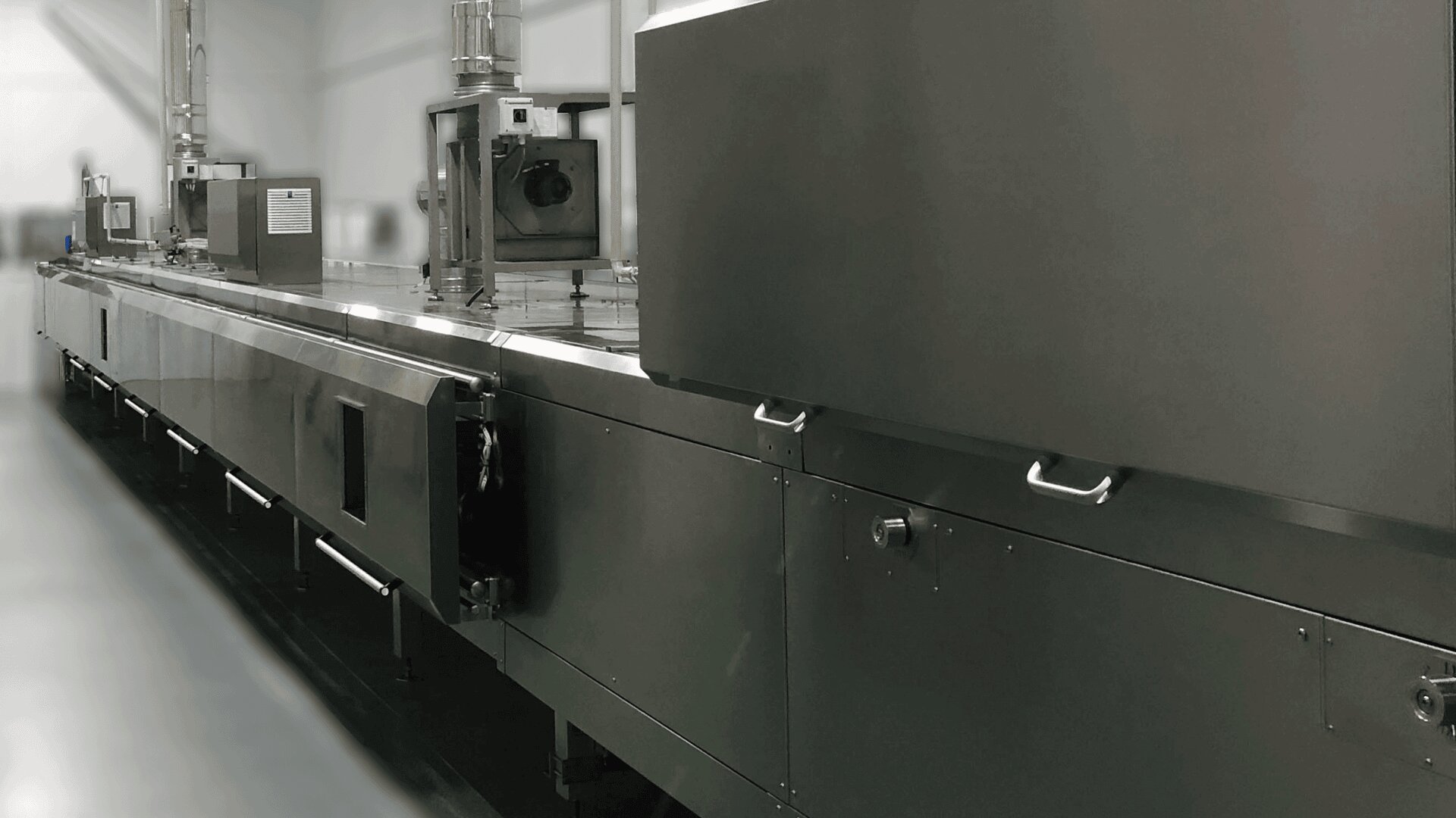 A long, industrial stainless steel food processing machine in a factory setting, with various compartments, control panels, and handles visible, situated in a clean, well-lit environment.