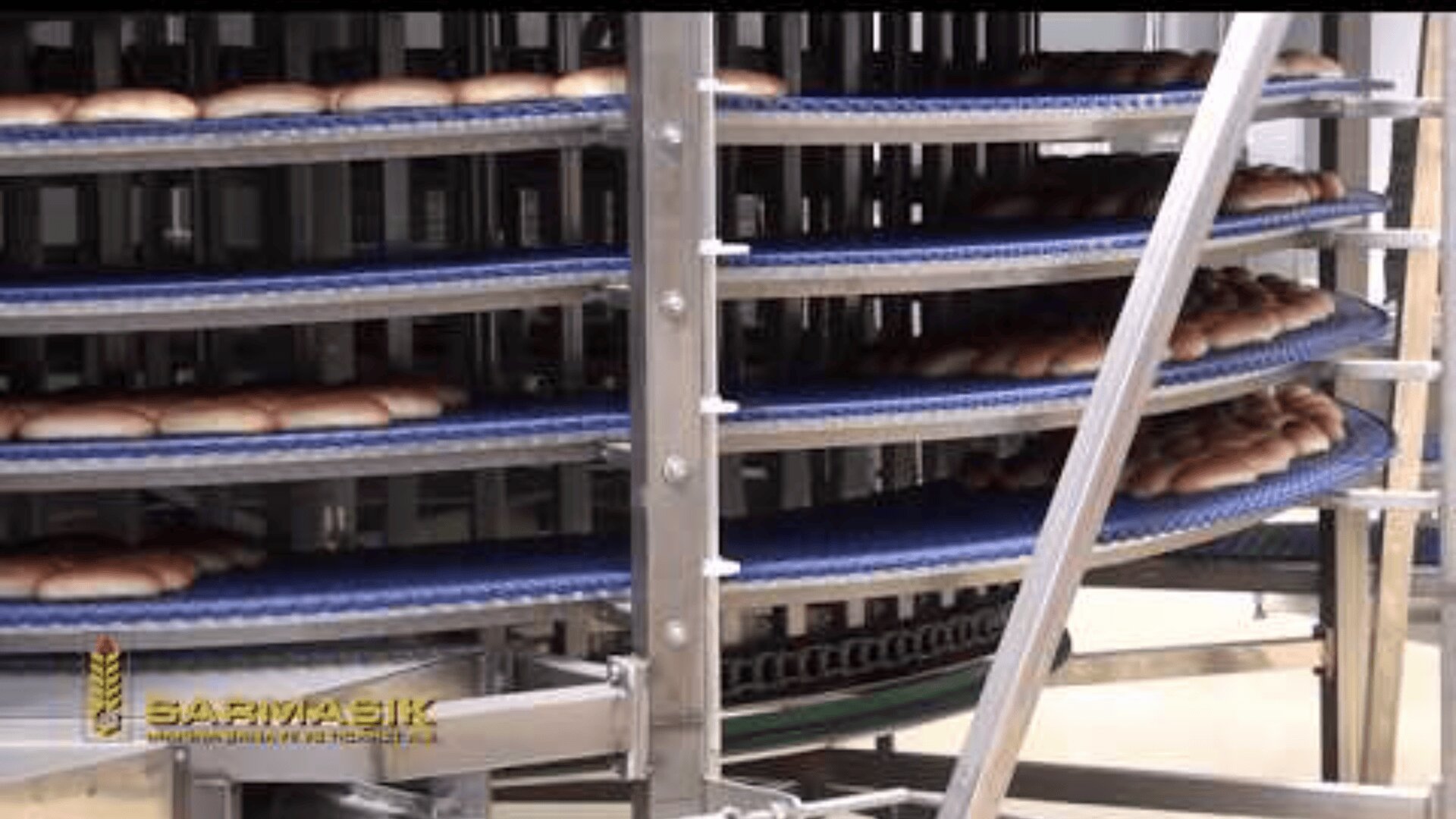 Close-up of a spiral conveyor system with multiple blue shelves, each holding rows of bread loaves, in an industrial bakery. A company logo, “SARIMASIK,” is visible in the lower left corner.