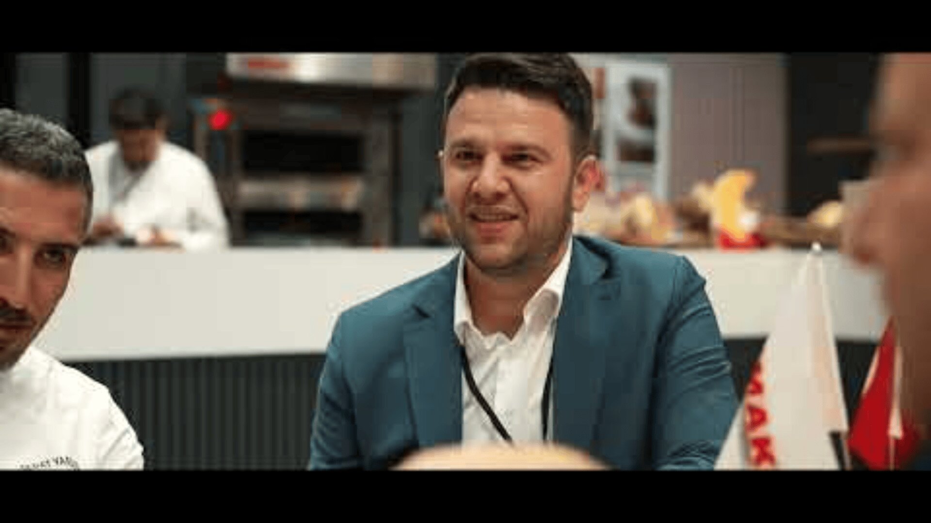 A man in a blue suit jacket sits at a table, smiling and talking with others in a modern indoor setting. People and flags are visible in the background.