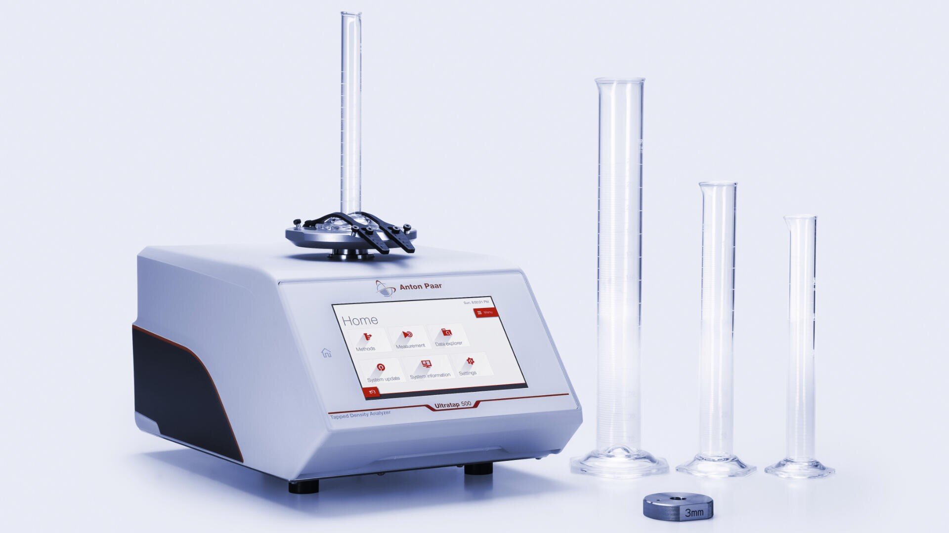 Auf weißem Hintergrund ist neben drei Messzylindern unterschiedlicher Größe und einem kleinen Metallgewicht ein Labordichtemessgerät mit digitalem Touchscreen-Display zu sehen.