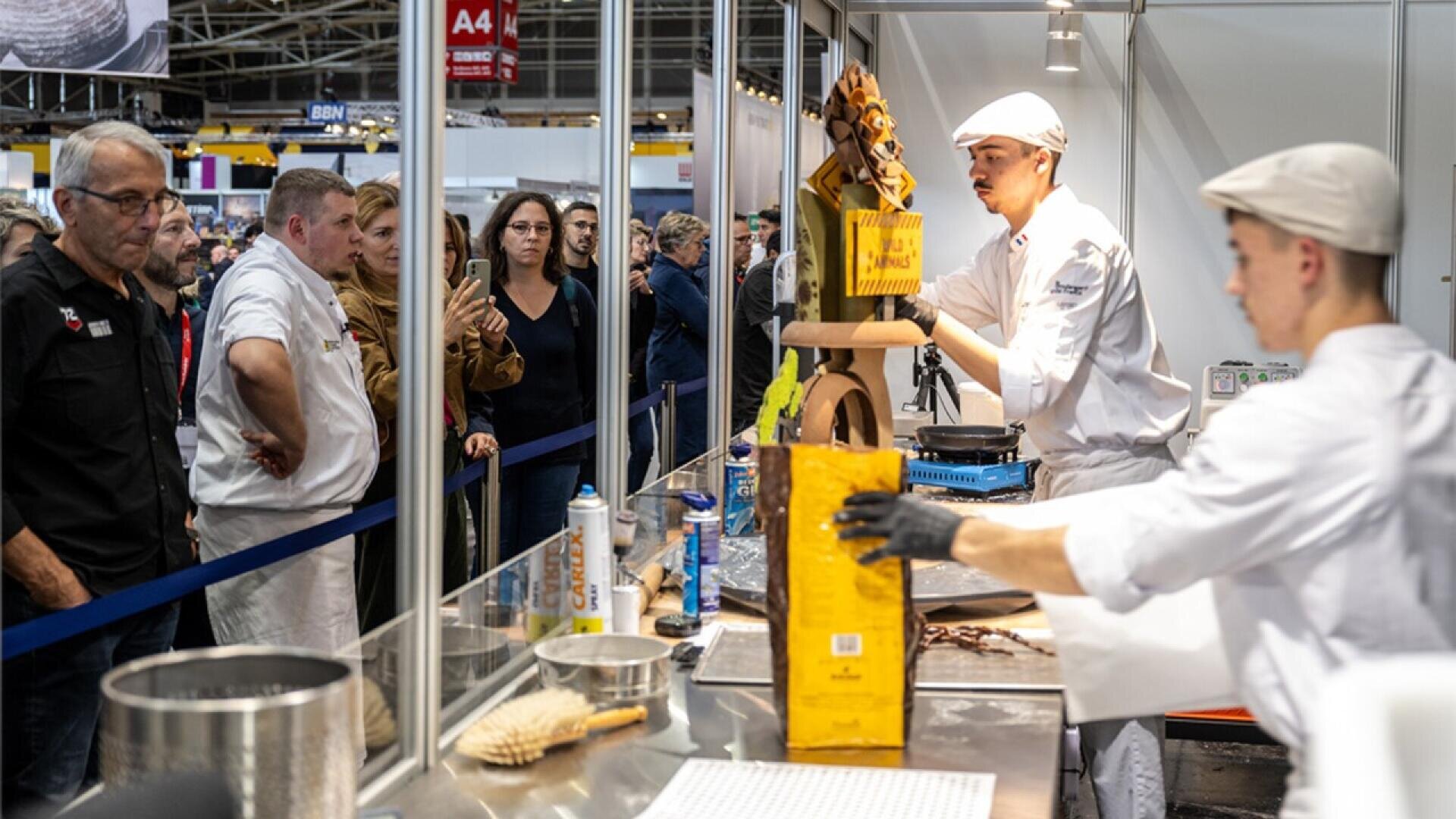 Two pastry chefs, dressed in white uniforms and hats, prepare chocolate desserts at a counter in front of an audience at a food event. One chef displays a chocolate creation while people watch attentively behind a barrier.