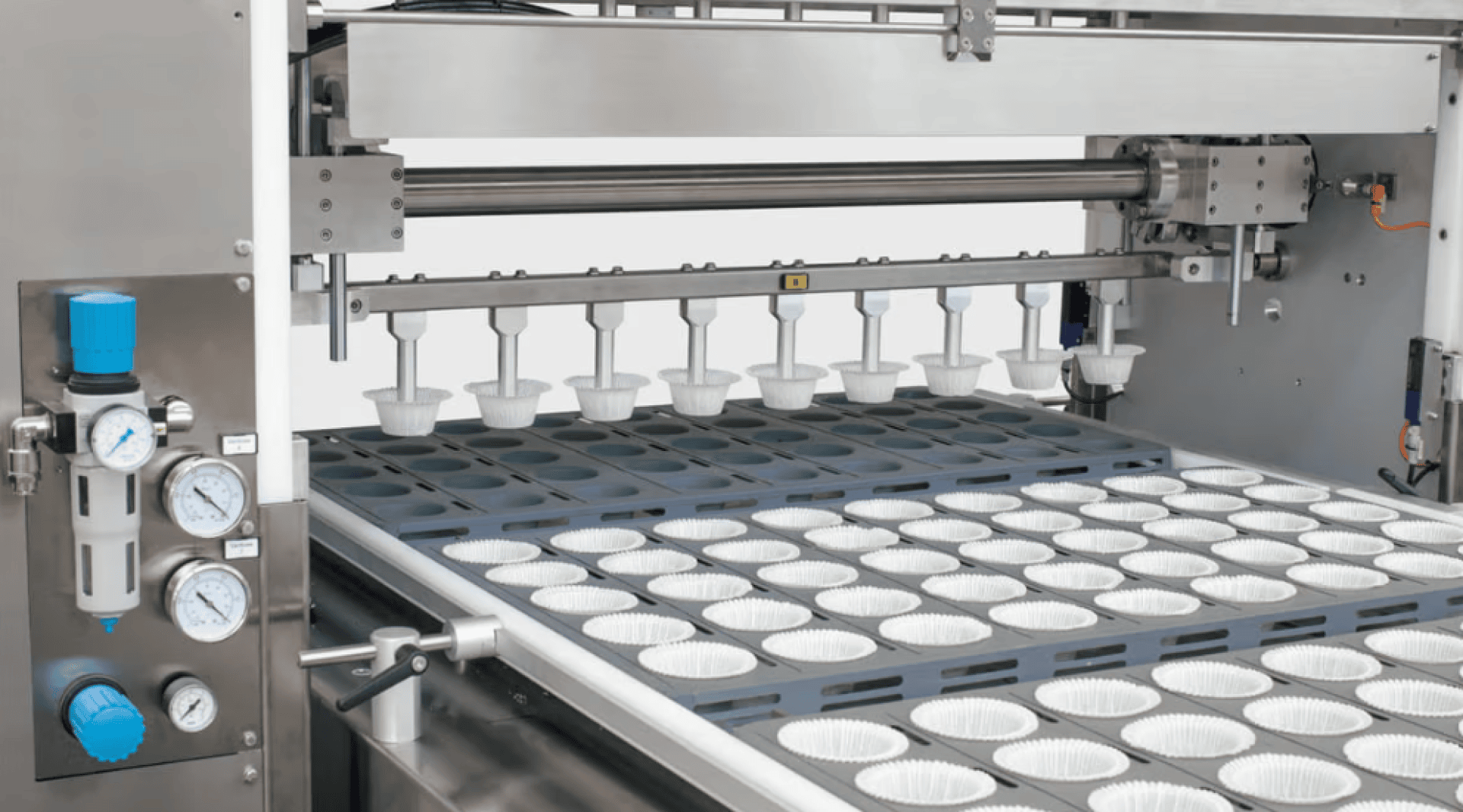 Nahaufnahme einer industriellen Bäckereimaschine, die Teig in Reihen von Muffinförmchen aus Papier auf einem Förderband verteilt, wobei auf der linken Seite verschiedene Bedienelemente und Anzeigen zu sehen sind.