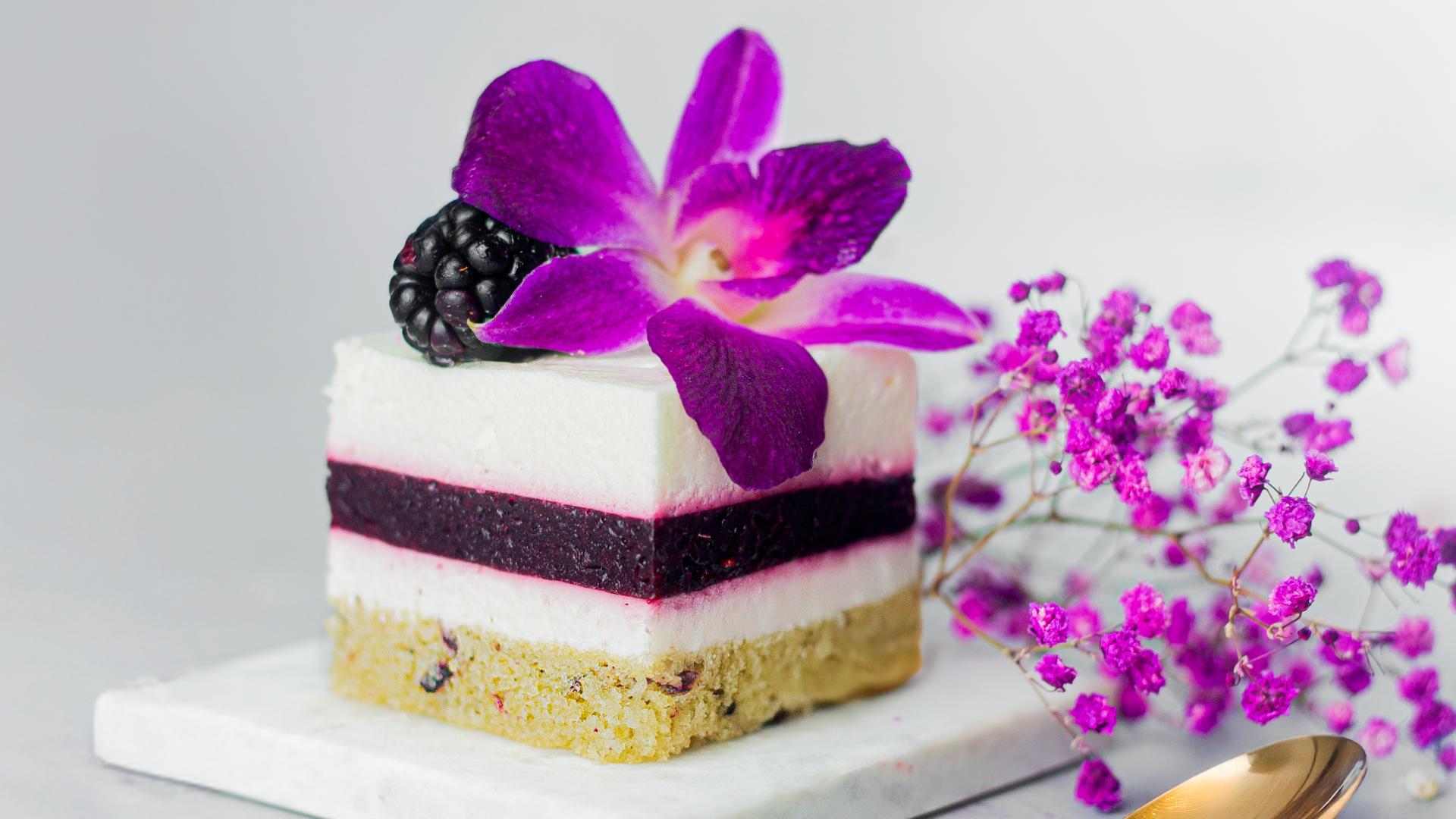 A slice of layered berry and cream dessert topped with a blackberry and a bright purple orchid, placed on a white marble slab, with small pink flowers and a gold spoon nearby.