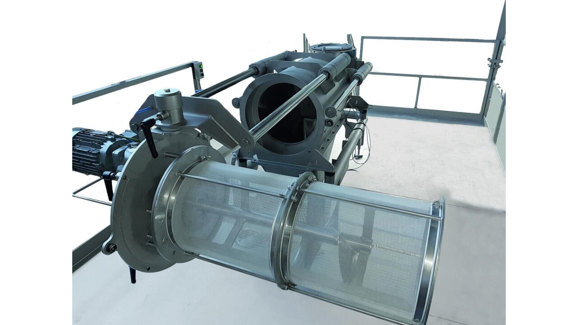 Industrial filtration machine with a cylindrical stainless steel mesh filter, pipes, and metal framework, situated on a raised platform with railing; the background is mostly white.