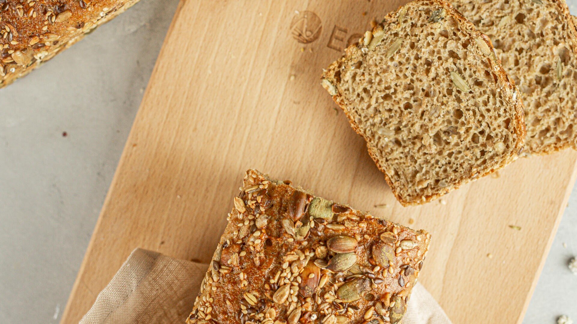 A loaf of multigrain bread and two slices rest on a wooden cutting board. The bread is topped with various seeds and grains. A beige cloth is partially visible underneath the loaf.