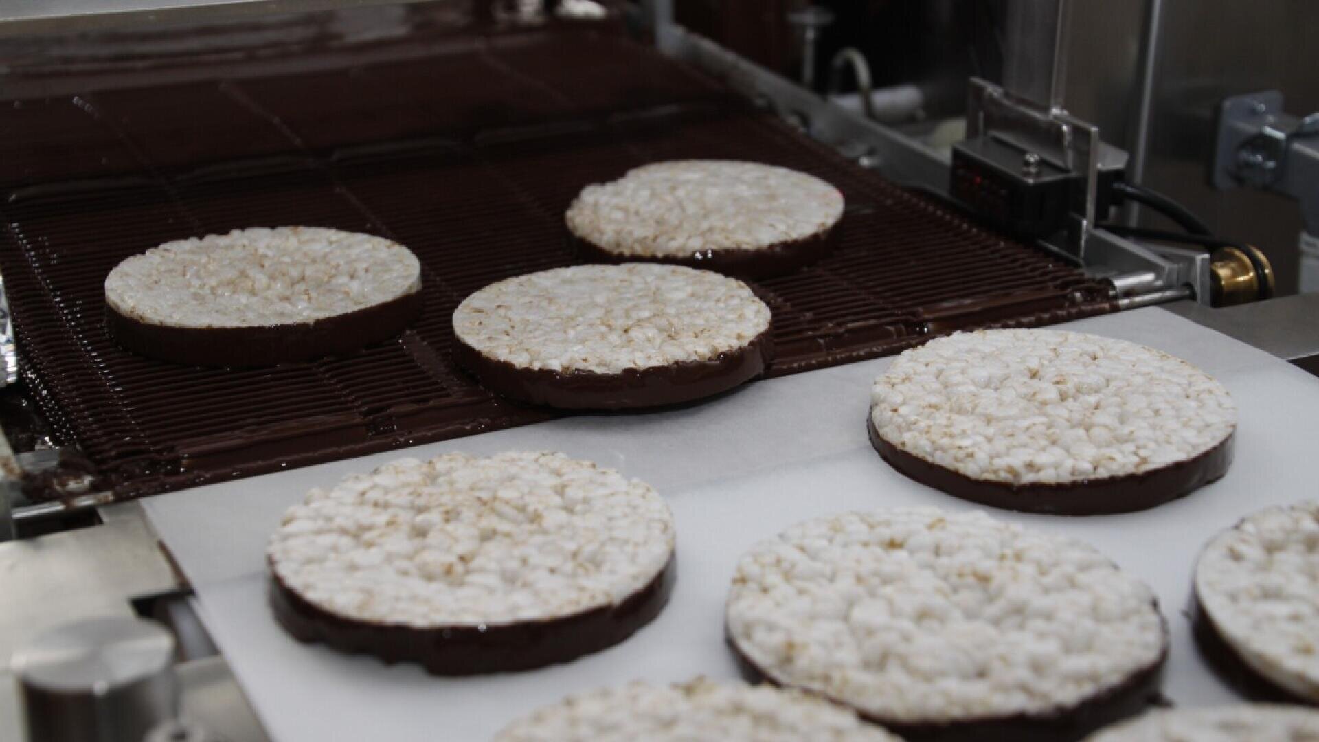 Large round rice cakes are being coated with chocolate on their bottom side as they move along a conveyor belt in a factory setting. Some cakes are on a metal grate, while others rest on white paper.