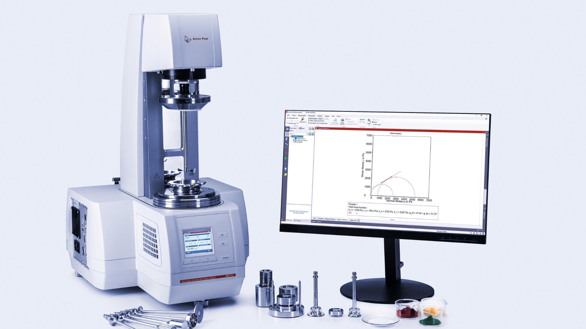 A laboratory rheometer next to a monitor displaying a graph, with several metal attachments and tools arranged on the table, all set against a light background.