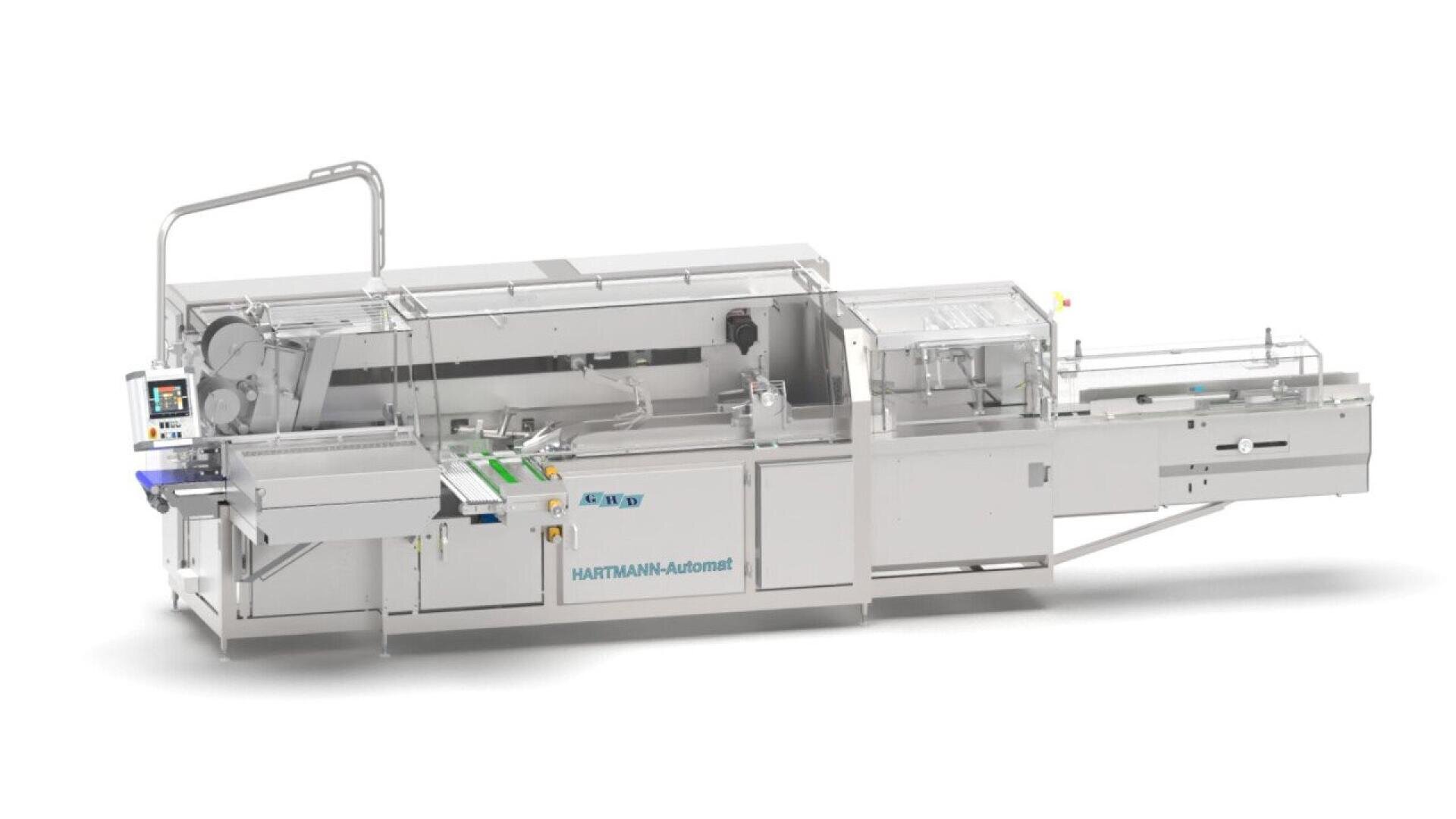 A large, industrial packaging machine labeled HARTMANN-Automat with conveyor belts, control panels, and compartments, designed for automated processing and packaging tasks in a factory setting.