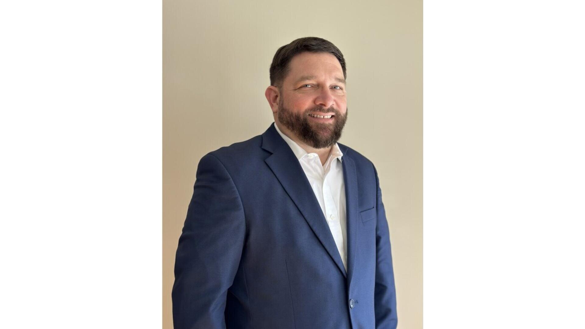 A man with short dark hair and a beard is wearing a dark blue suit jacket and a white collared shirt, standing and smiling in front of a plain light-colored background.