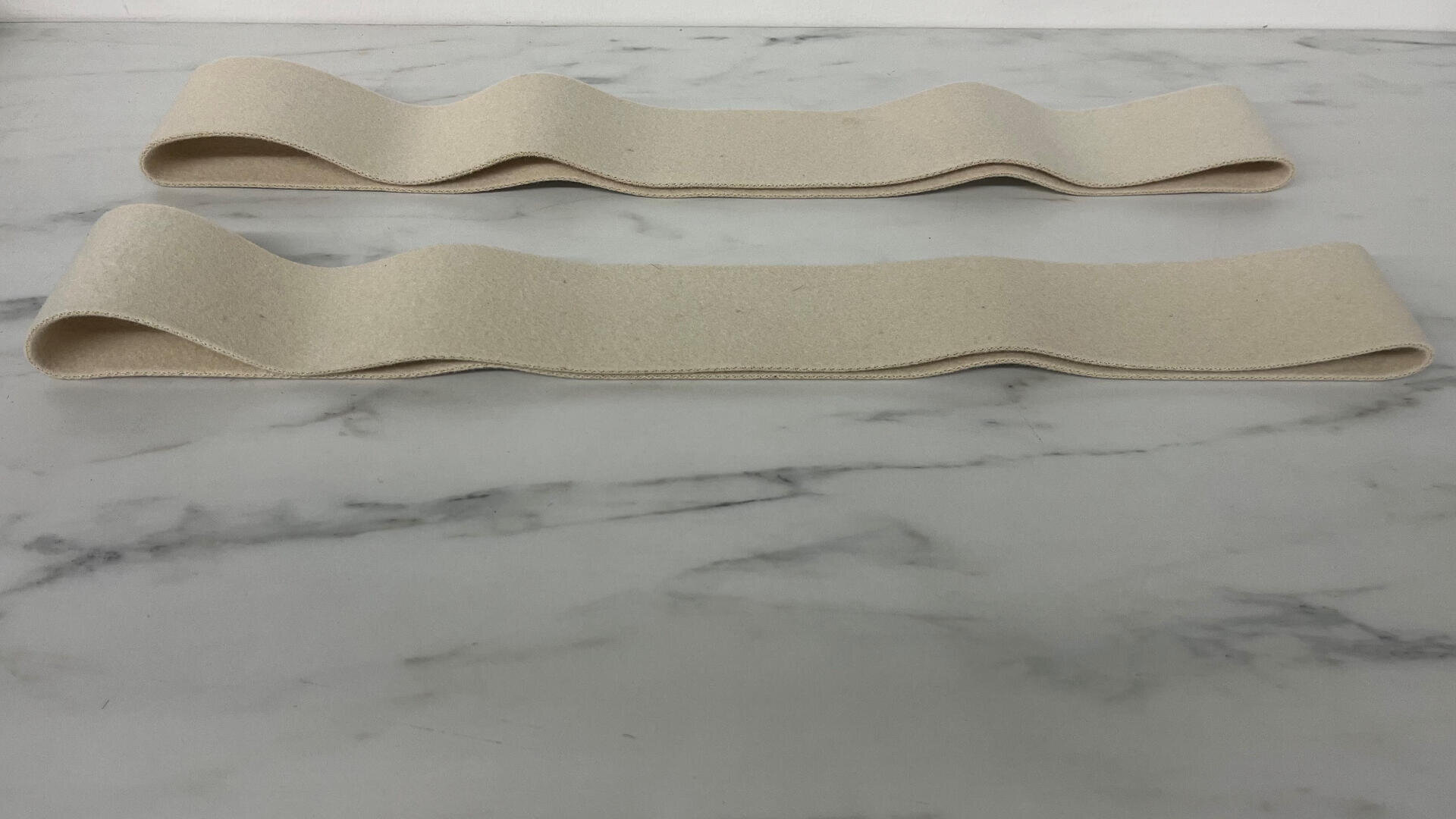 Zwei beigefarbene, flache Stoffschleifen mit gewellten Kanten liegen parallel auf einer weißen Marmorfläche.