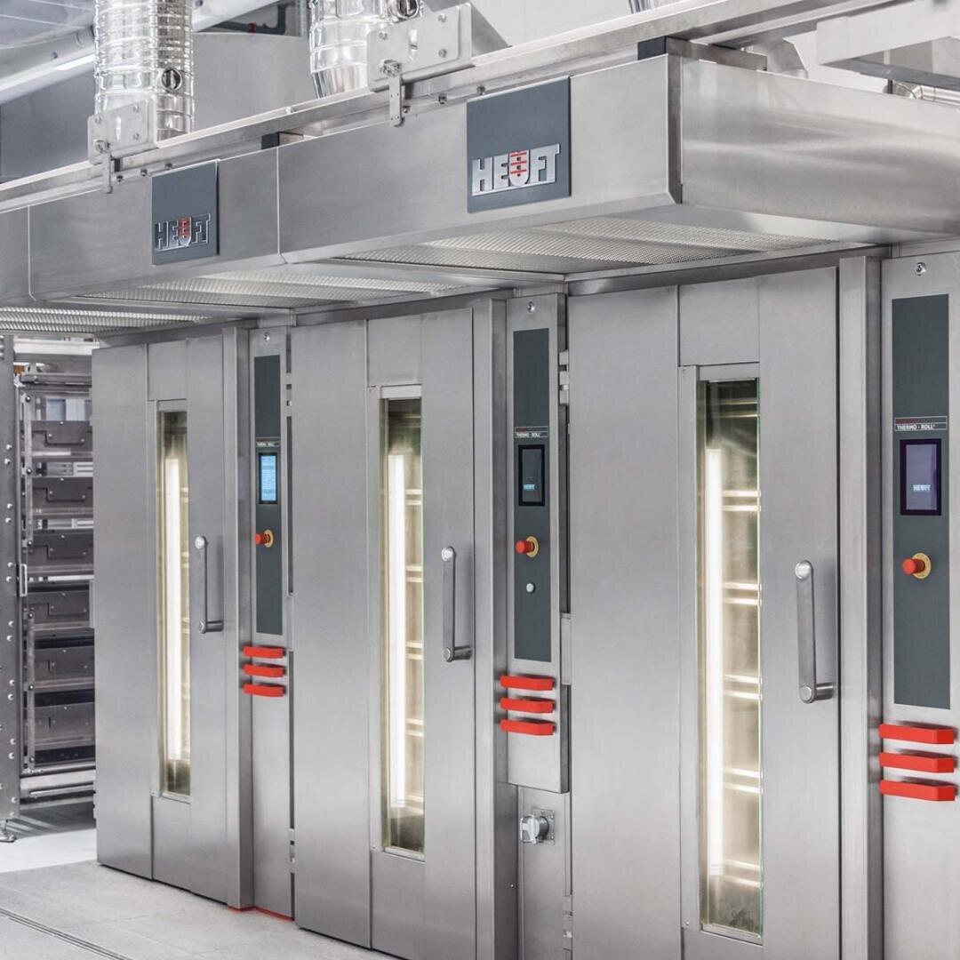 Eine Reihe großer Industrieöfen aus Edelstahl mit vertikalen Glasfenstern und digitalen Bedienfeldern in einer modernen Großküche oder Bäckerei.