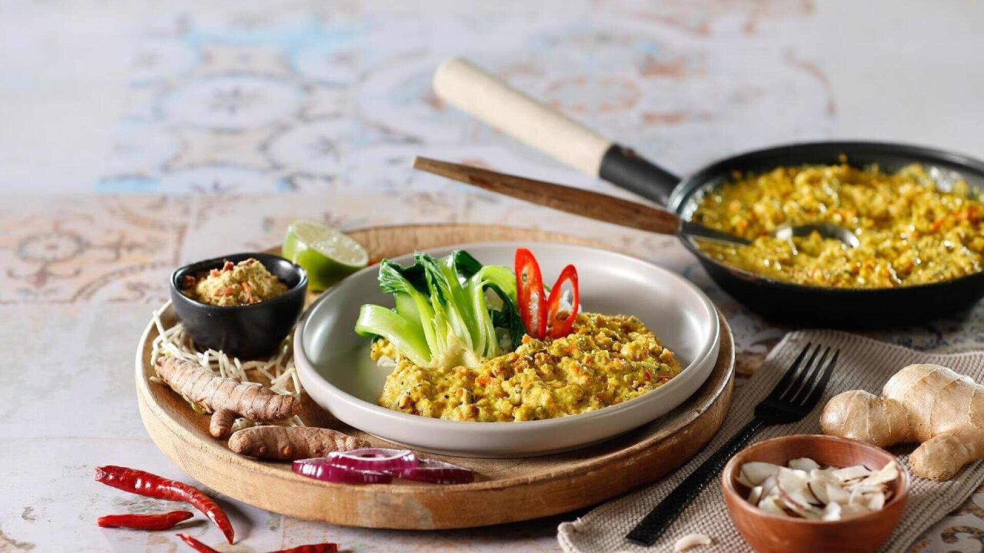 A plate of golden-colored rice, garnished with sliced red chili and greens, sits on a round platter with fresh ingredients like ginger, turmeric, chili, and lime; a skillet of rice is in the background.