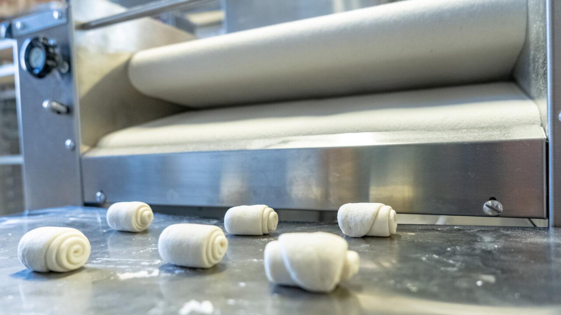 Kleine gerollte Teigstücke liegen auf einer Metallfläche vor einer kommerziellen Teigausrollmaschine in einer Bäckerei.