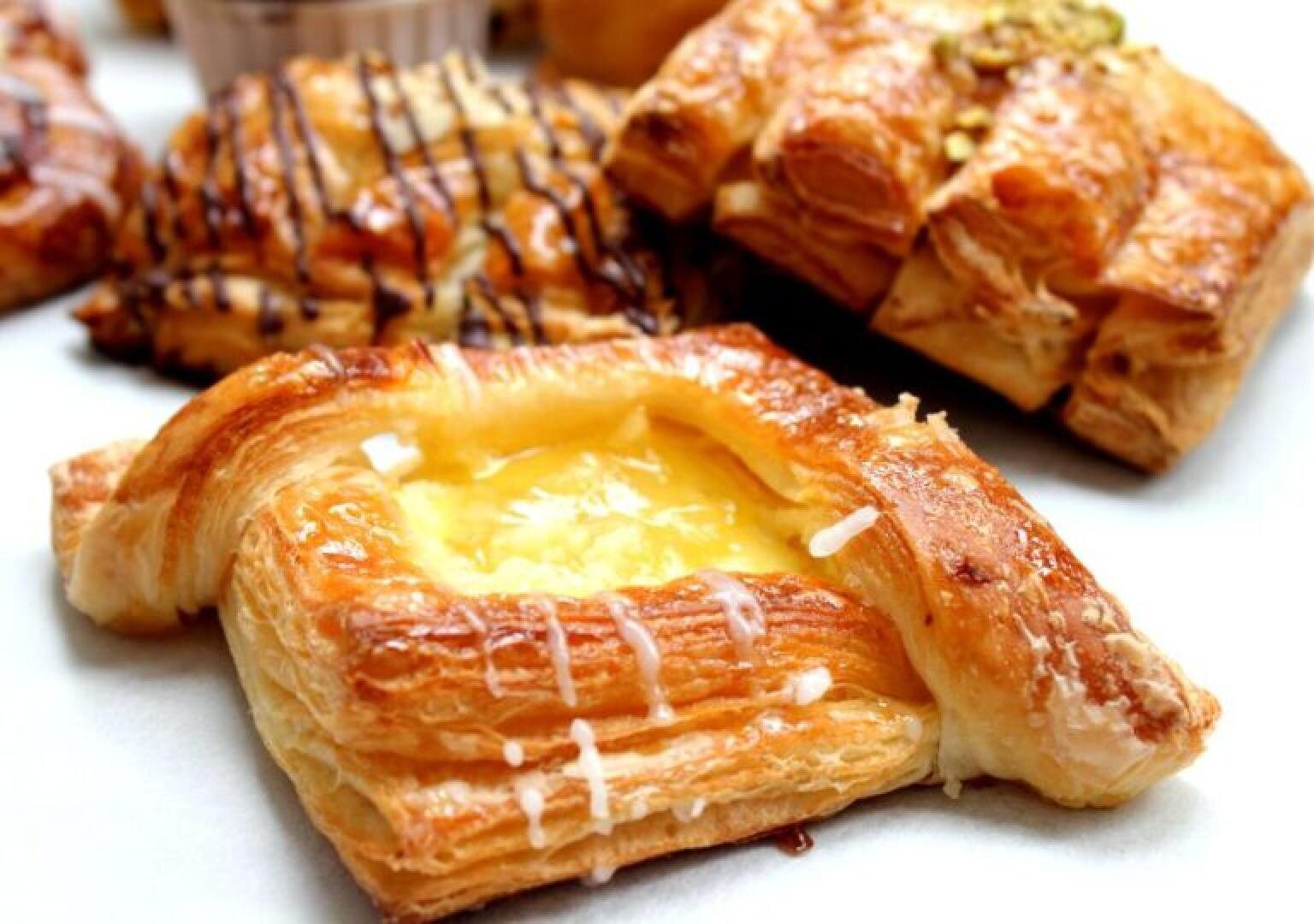 A close-up of assorted pastries, including a flaky Danish with a creamy yellow custard center and a drizzle of icing, surrounded by other pastries with chocolate drizzle and nut toppings.