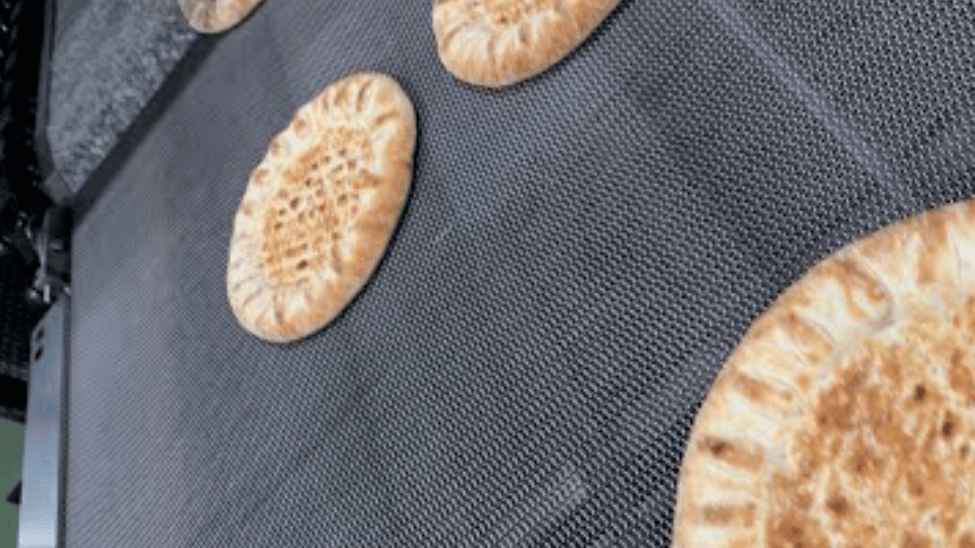 Vier runde, goldbraune Fladenbrote backen auf einem Förderband in einem Industrieofen.