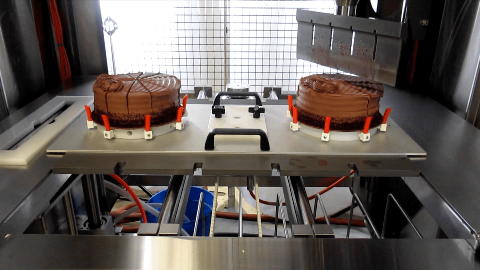 Two chocolate cakes sit on a metal tray inside an industrial slicing machine, with red clamps holding them in place and a grid barrier in the background.
