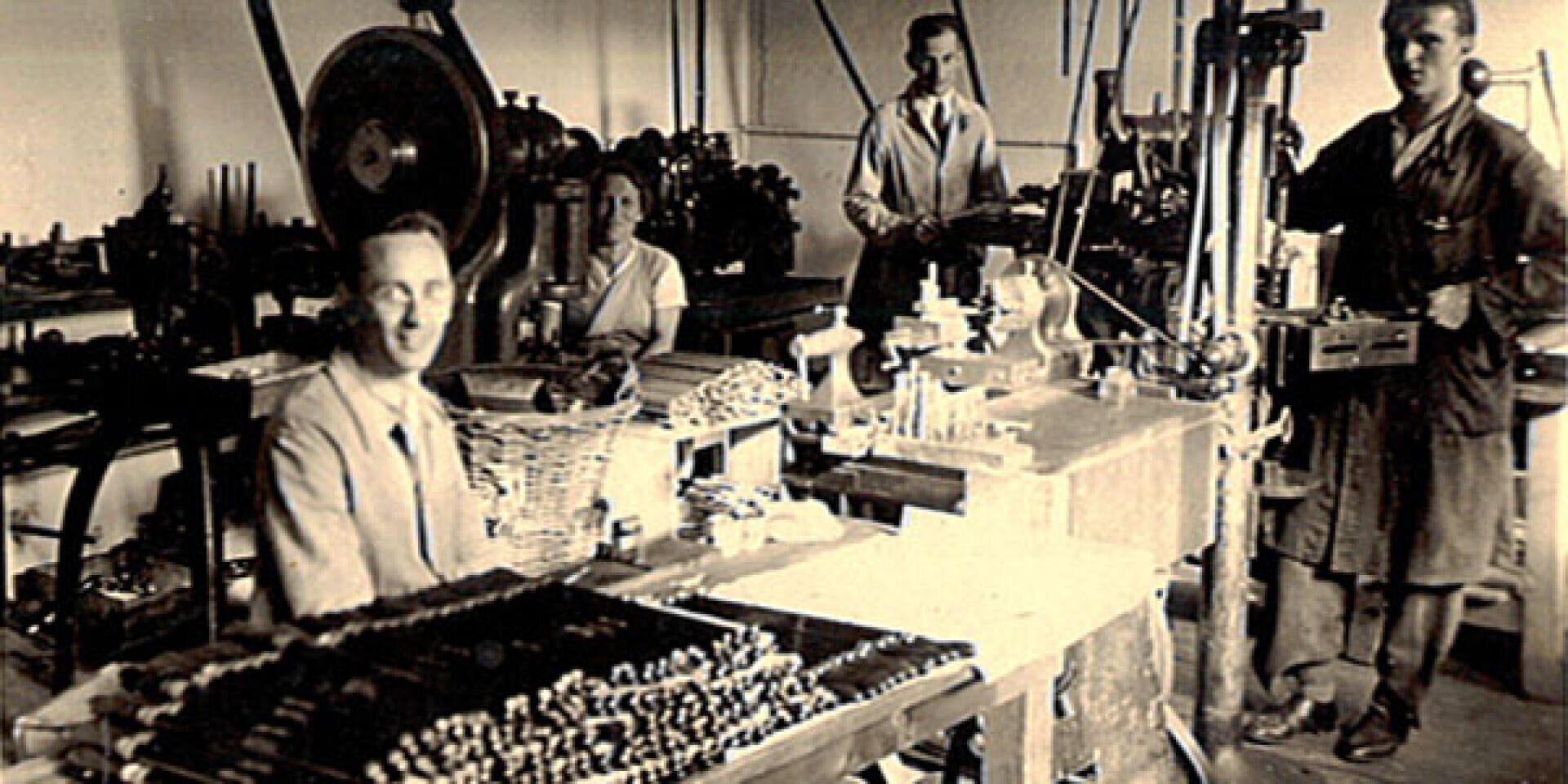 Four people work in a vintage factory workshop with various mechanical equipment and tools on tables. The room is cluttered, and the workers wear formal and industrial attire, suggesting an early to mid-20th century setting.