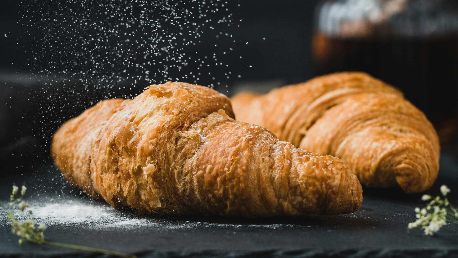 Two golden, flaky croissants sit on a dark surface as powdered sugar is sprinkled over them, creating a light dusting. The background is blurred, highlighting the croissants’ crisp, layered texture.