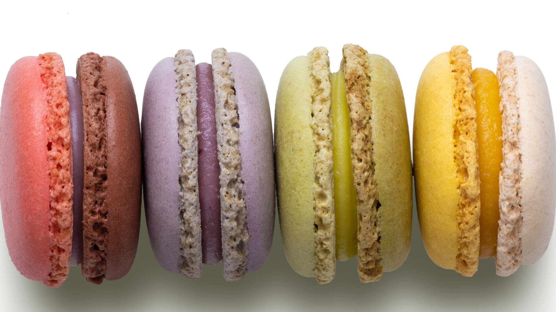 Fünf bunte Macarons in einer Reihe mit roten, braunen, lila, grünen, gelben und cremefarbenen Tönen auf weißem Hintergrund.