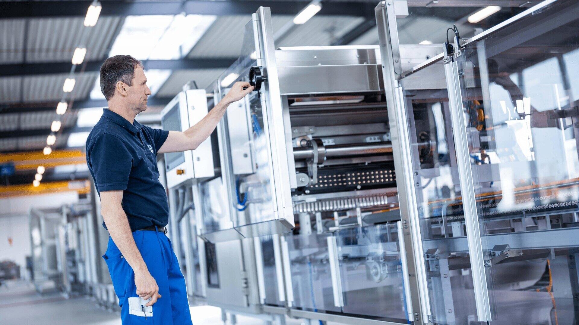 Ein Mann in einem marineblauen Hemd und einer blauen Hose bedient eine große, moderne Industriemaschine in einer hell erleuchteten Fabrikhalle. Mit seiner rechten Hand stellt er ein Bauteil an der Maschine ein.