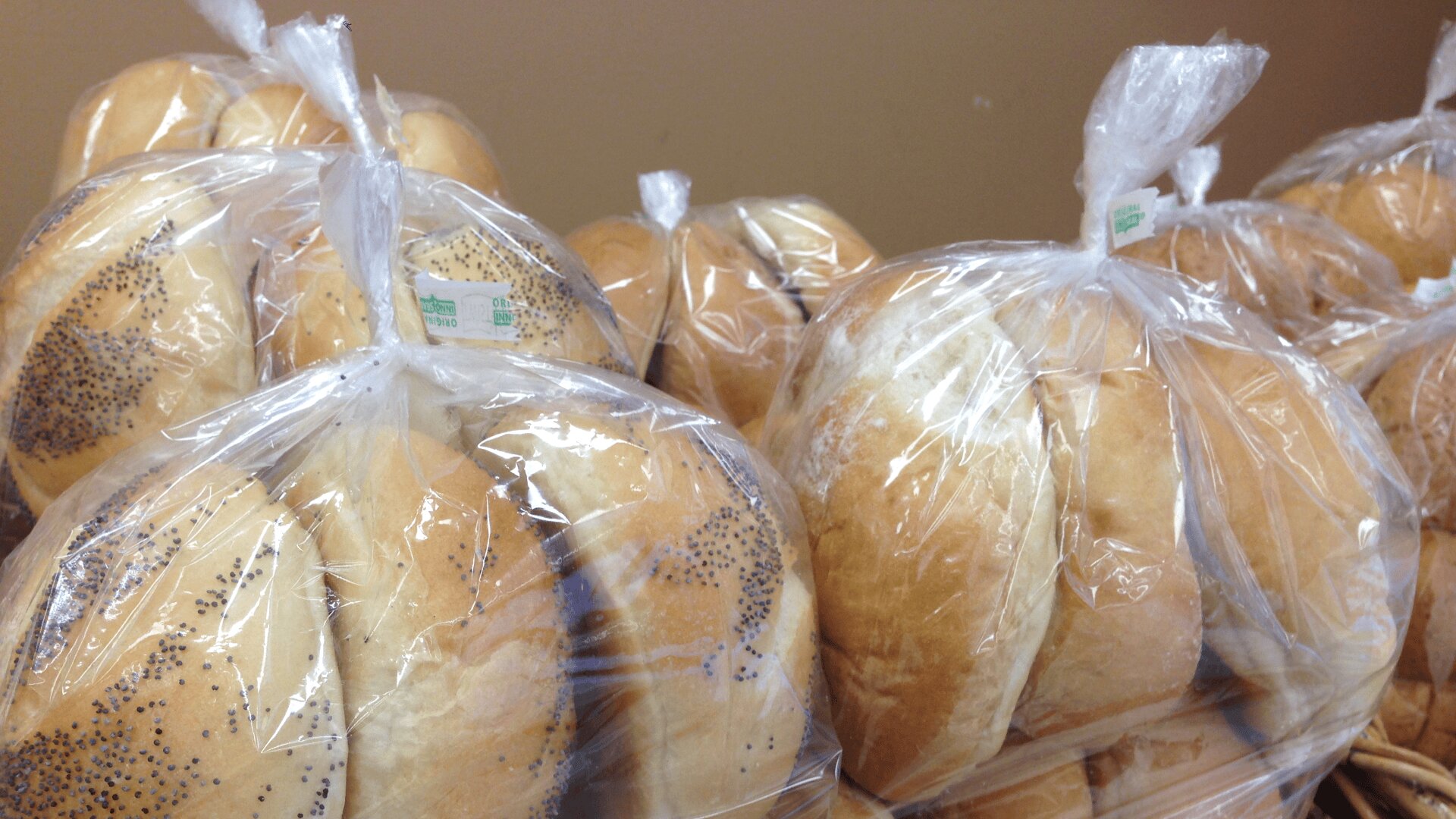 Several packages of hamburger buns, some with poppy seeds, are wrapped in clear plastic bags and displayed against a plain beige background.