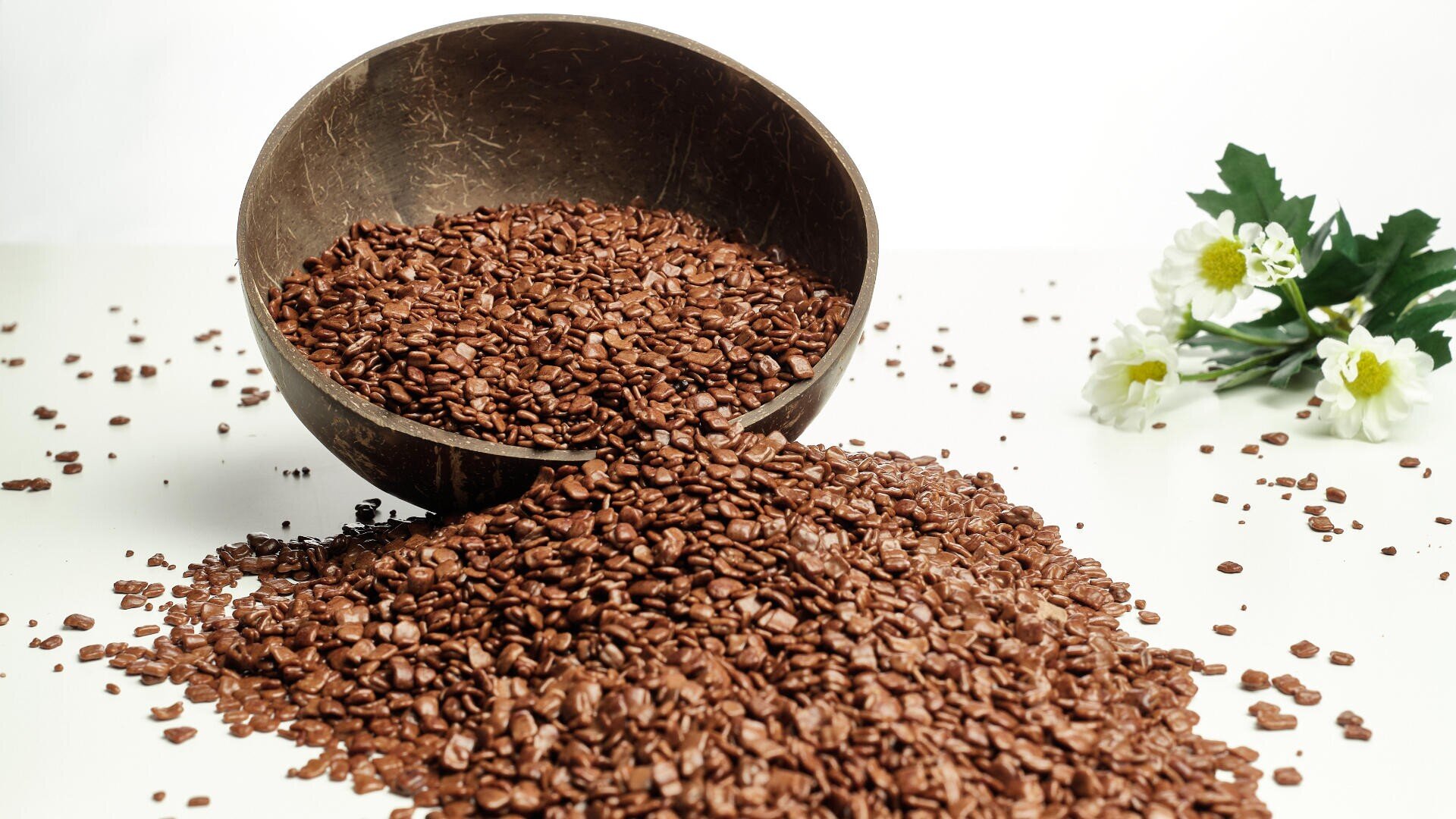 A brown coconut bowl tipped over, spilling a large pile of flax seeds onto a white surface, with white and yellow flowers in the background.