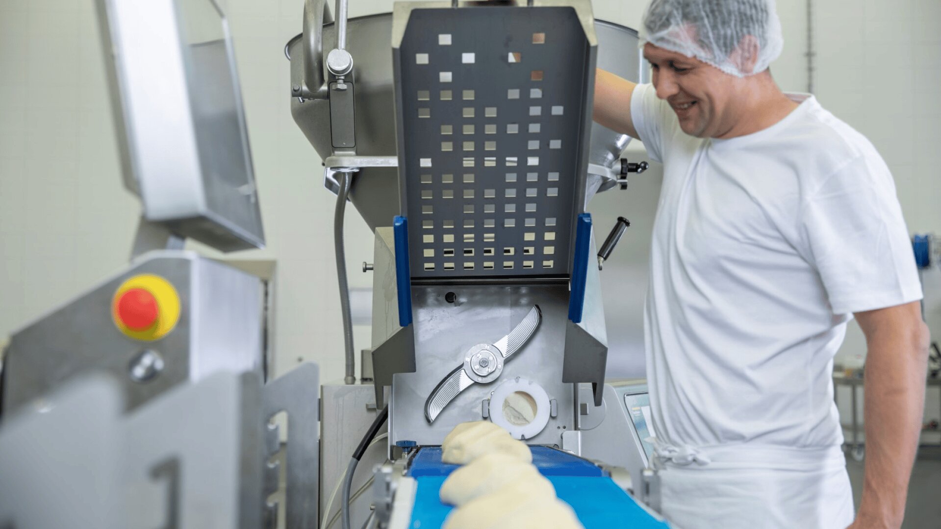 A worker in a white uniform and hairnet smiles while operating a machine that processes dough pieces on a blue conveyor belt in a food production facility.