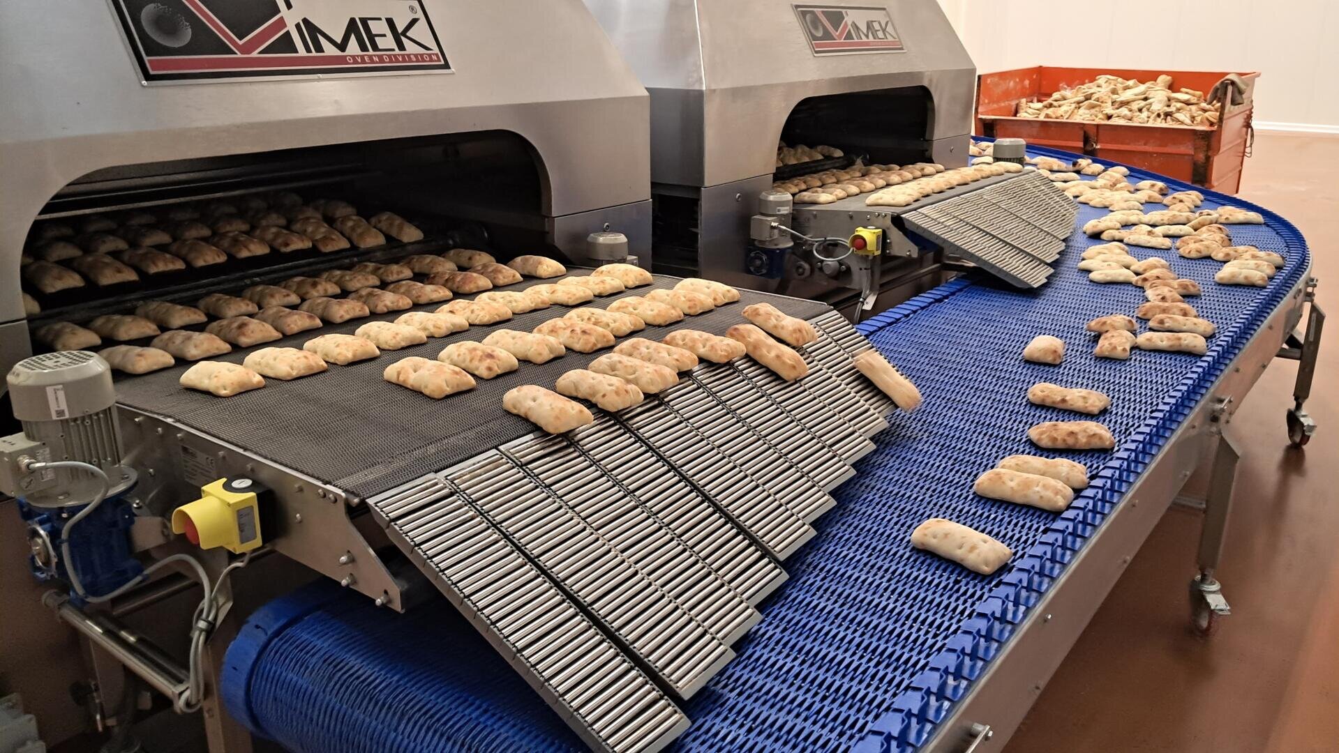 Rows of rectangular bread rolls move on a blue conveyor belt from industrial ovens in a bakery, with baked goods cooling and piling up on the conveyor system.