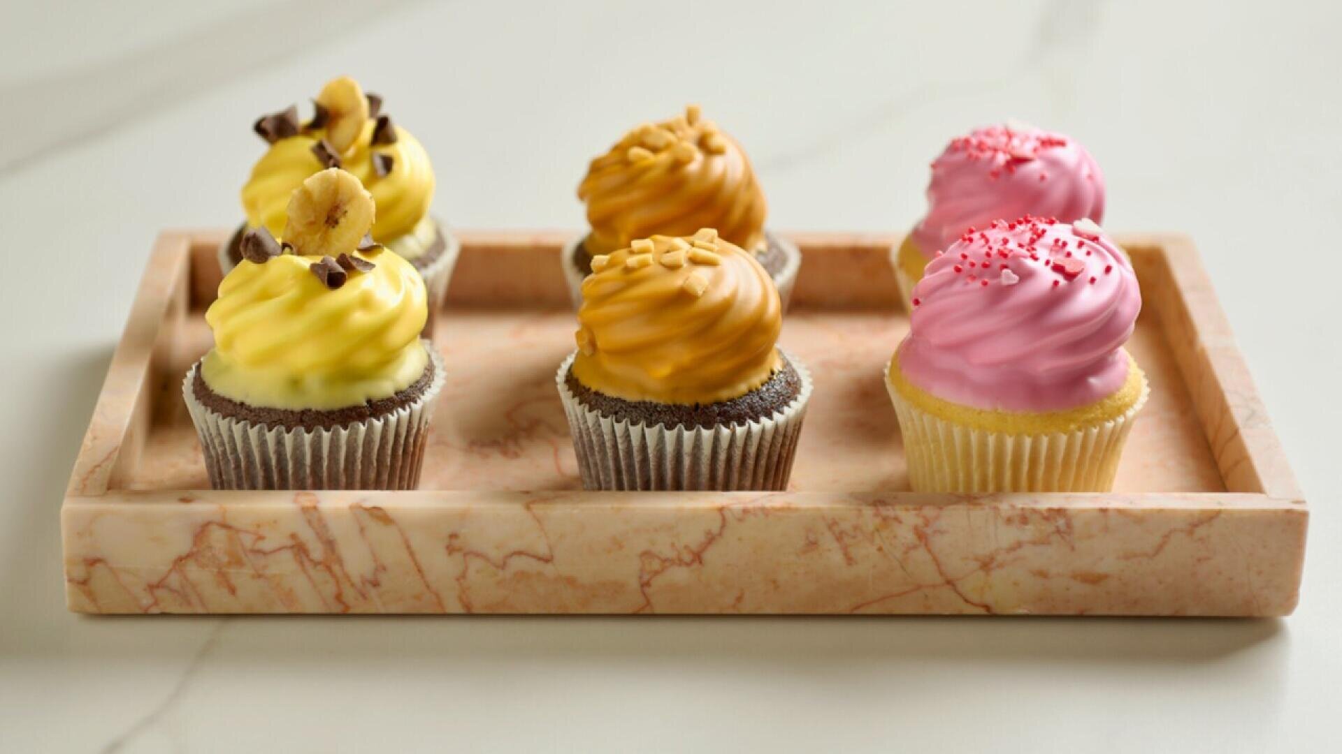 Auf einem Marmortablett liegen sechs Cupcakes: zwei mit gelbem Zuckerguss und Schokoladendekoration, zwei mit karamellfarbenem Zuckerguss und zwei mit rosa Zuckerguss und roten Streuseln.