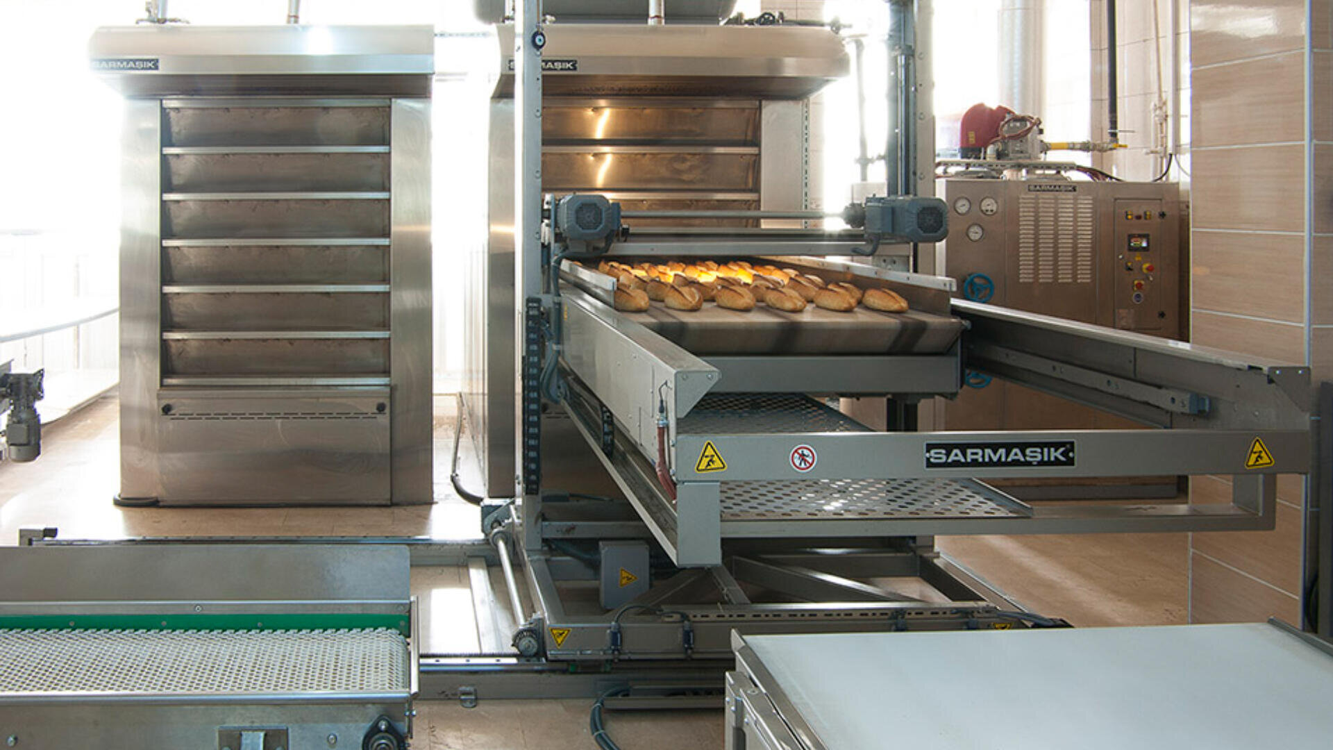 Eine kommerzielle Brotbackmaschine mit Blechen mit frisch gebackenen Broten, die auf einem Förderband in einer modernen industriellen Bäckerei herauskommen.