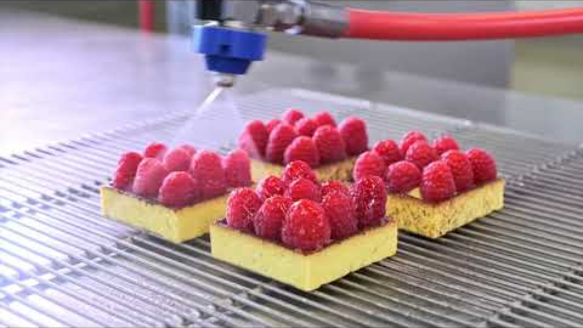 Four square raspberry desserts are sprayed with a clear glaze by a machine nozzle on a metal cooling rack. The glaze gives the raspberries a shiny appearance.