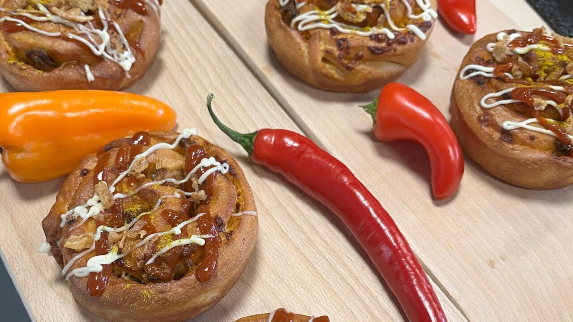 Swirled rolls topped with sauces and fried onions are arranged on a wooden board alongside whole orange and red chili peppers.