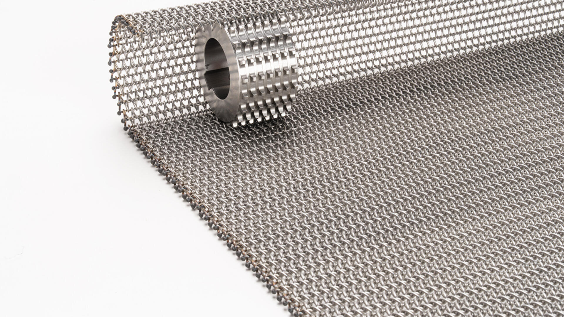 Eine Nahaufnahme einer Rolle aus Metallgeflecht oder Drahtgewebe mit einem zylindrischen Metallstück im Inneren der Rolle, dargestellt auf einer weißen Oberfläche.