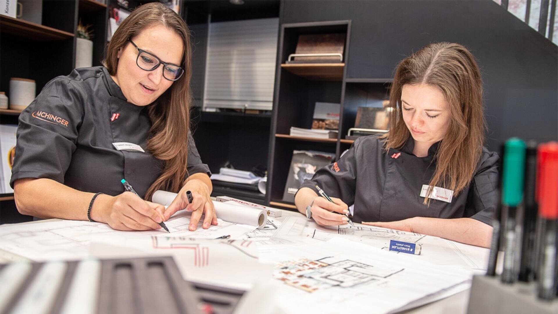 Two women in dark uniforms sit at a desk covered with architectural drawings and papers, focused on writing and sketching plans. Shelves with office supplies are visible in the background.