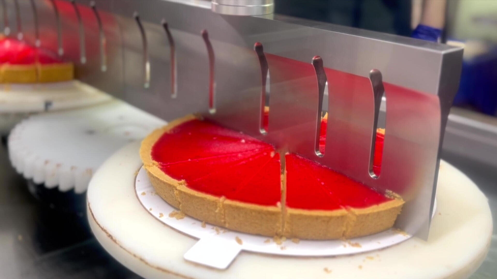 A machine slices a bright red pie with a graham cracker crust into even pieces on a rotating white platform.