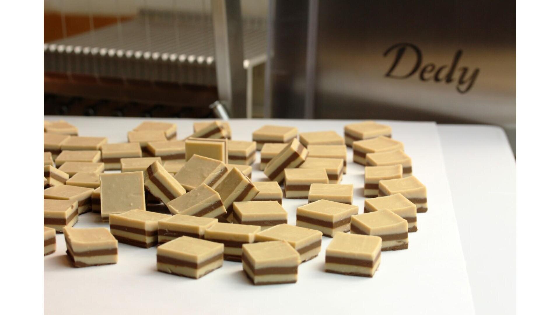 A pile of square, layered chocolates with light and dark stripes sits on a white surface. In the background, theres stainless steel equipment with the word Dedy engraved on it.