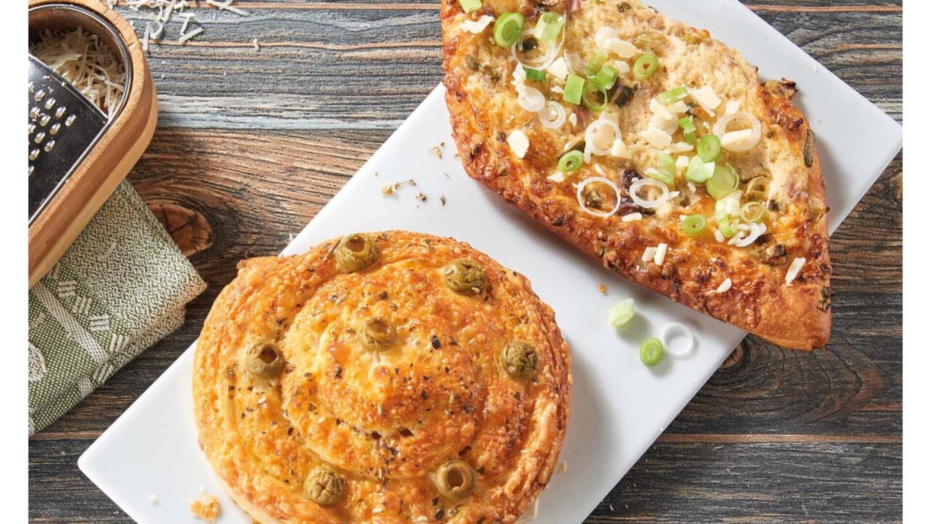 A rectangular plate with a round cheesy pastry topped with green olives and a wedge-shaped bread topped with chopped green onions and cheese, set on a wooden surface next to a cheese grater.