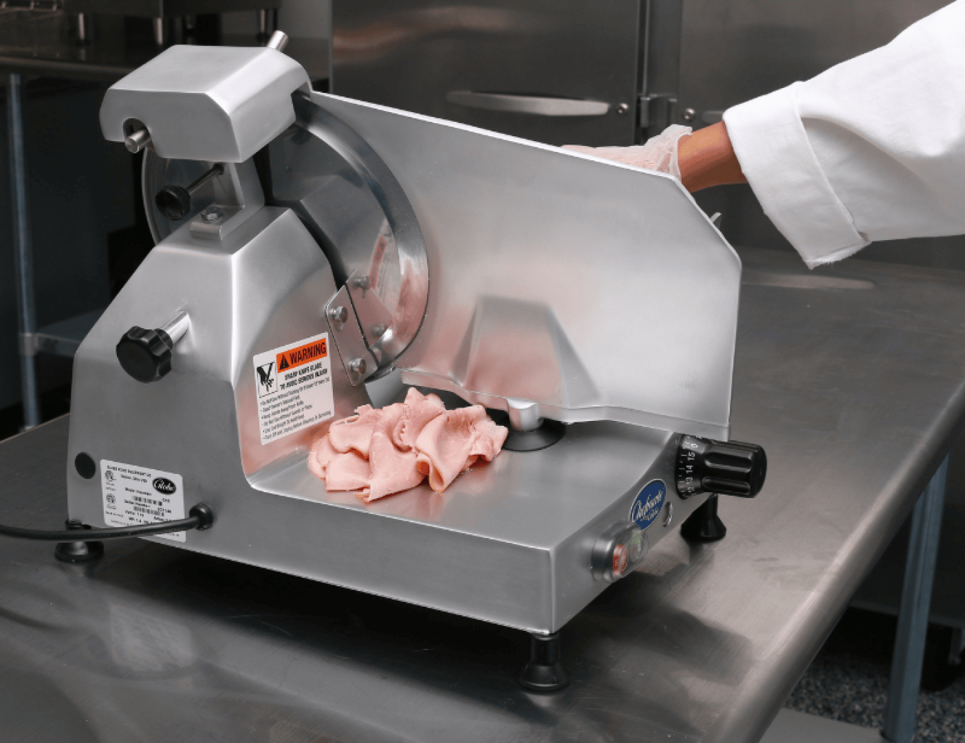 Eine Person in weißem Kittel und mit Handschuhen schneidet mit einer handelsüblichen Aufschnittmaschine auf einer Arbeitsplatte aus Edelstahl in einer Küche Feinkostfleisch auf.