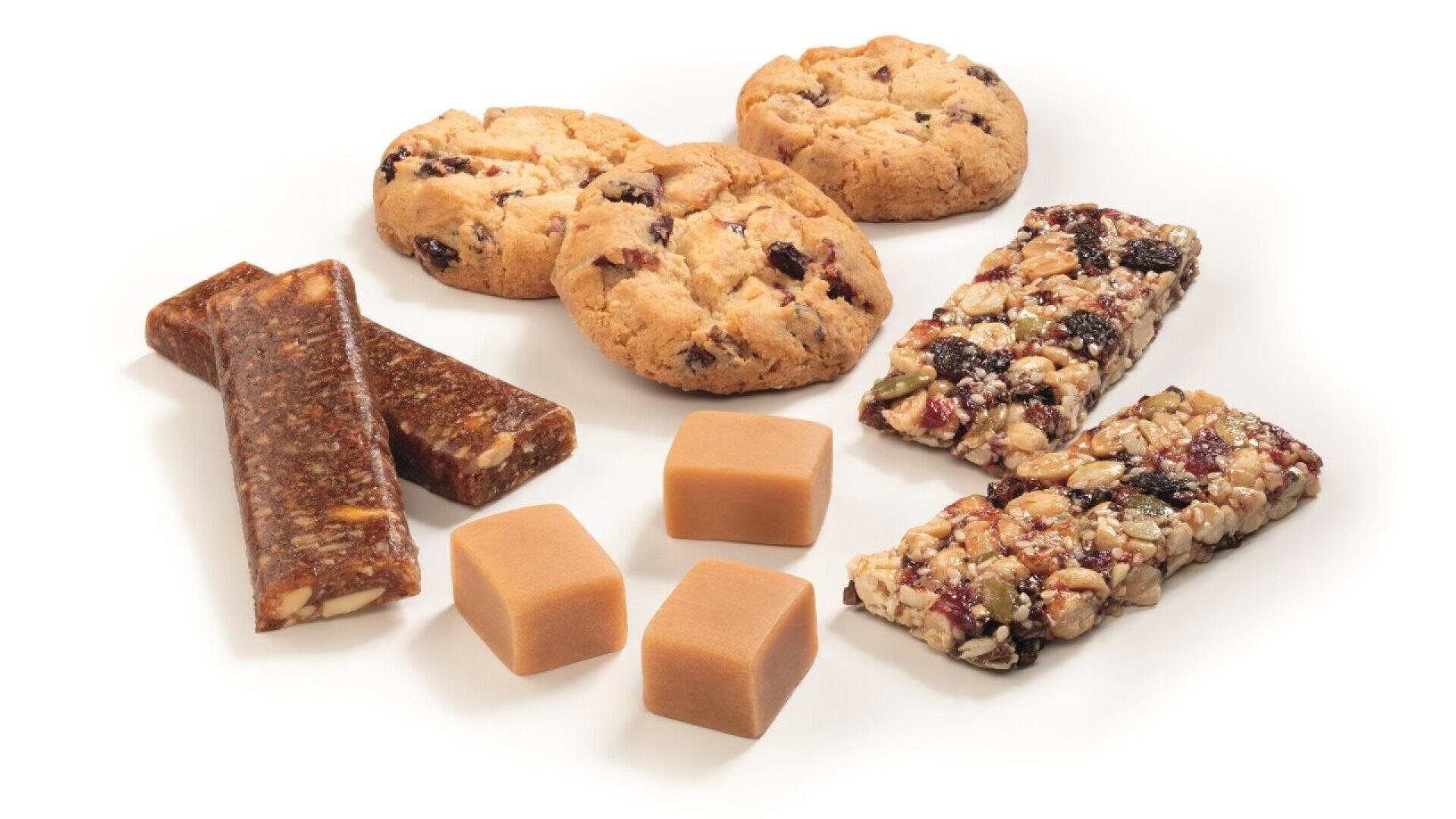 An assortment of snacks including three round cookies, two chewy granola bars, two rectangular nut bars, and three caramel cubes, all arranged on a white background.