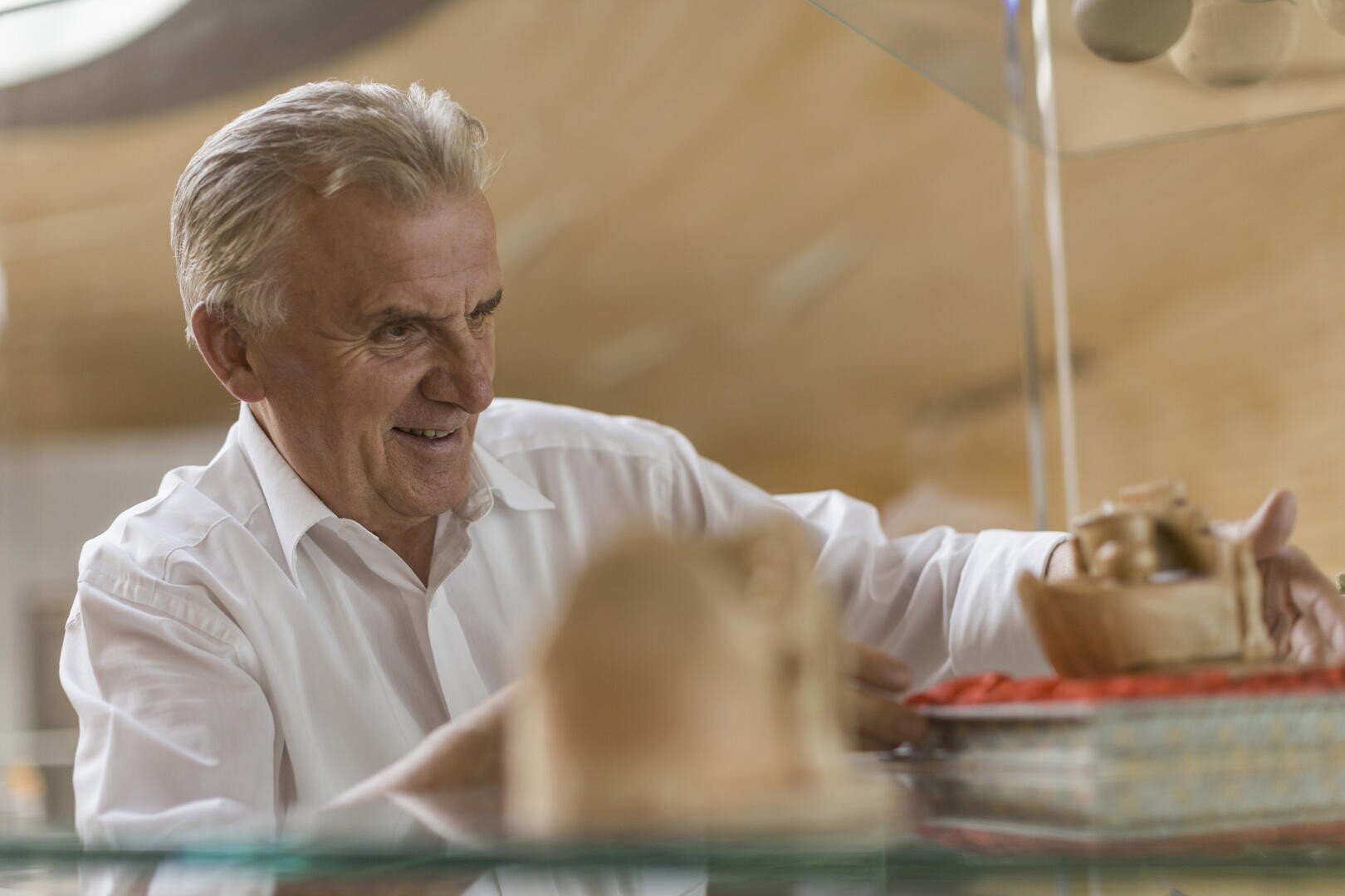 Ein älterer Mann in einem weißen Hemd lächelt, während er in einem warm beleuchteten Raum ein Holzobjekt auf einem Glasregal arrangiert. Im Vorder- und Hintergrund sind weitere Holzgegenstände zu sehen.
