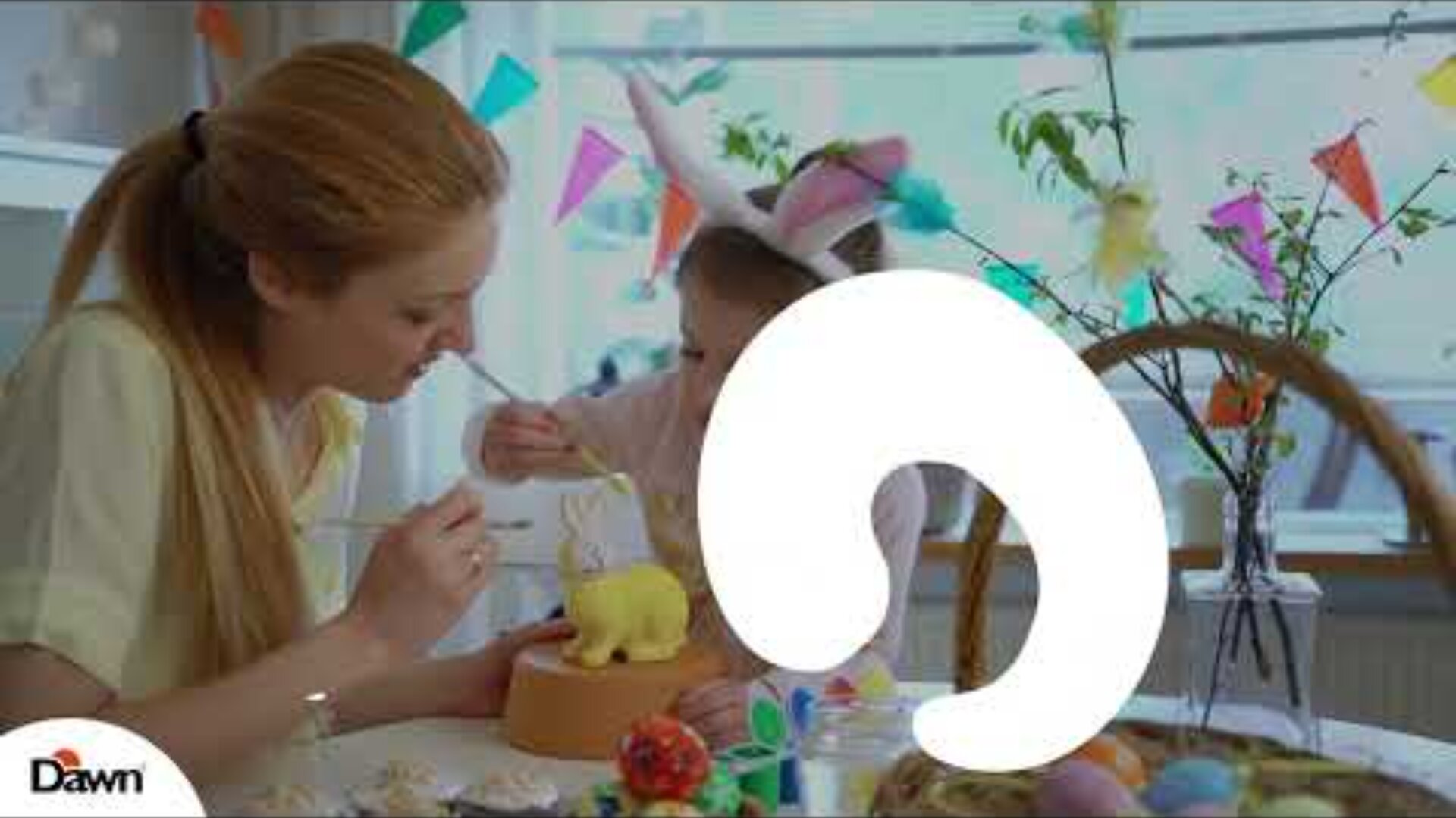 A woman and a child wearing bunny ears decorate an Easter cake at a festive table with colorful eggs and decorations. Bright party flags hang in the background. Dawn logo appears in the bottom left corner.