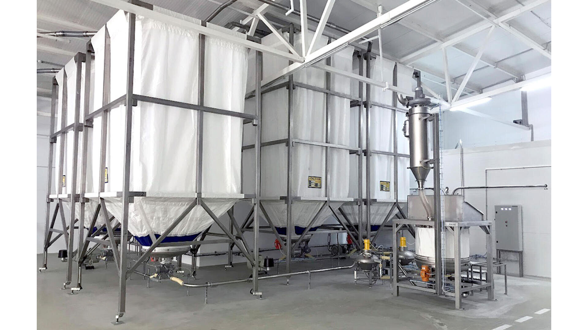 Large industrial silos with white cladding and metal frames stand in a bright, clean facility. Pipes, valves, a control panel and other processing equipment are visible around the machines.