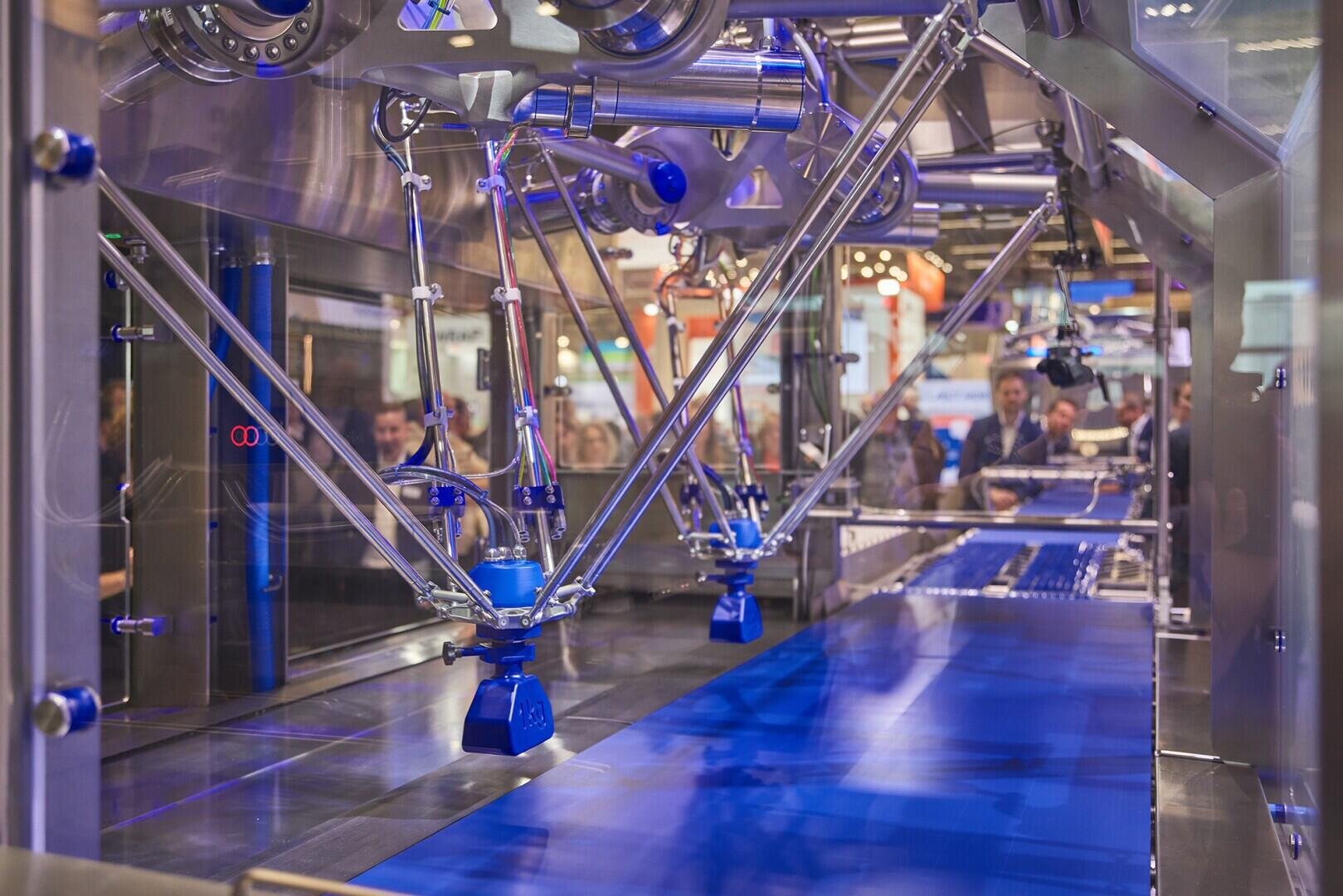 A robotic arm system with multiple metal arms and blue suction cups operates above a blue conveyor belt inside a factory, while people observe through a glass panel in the background.