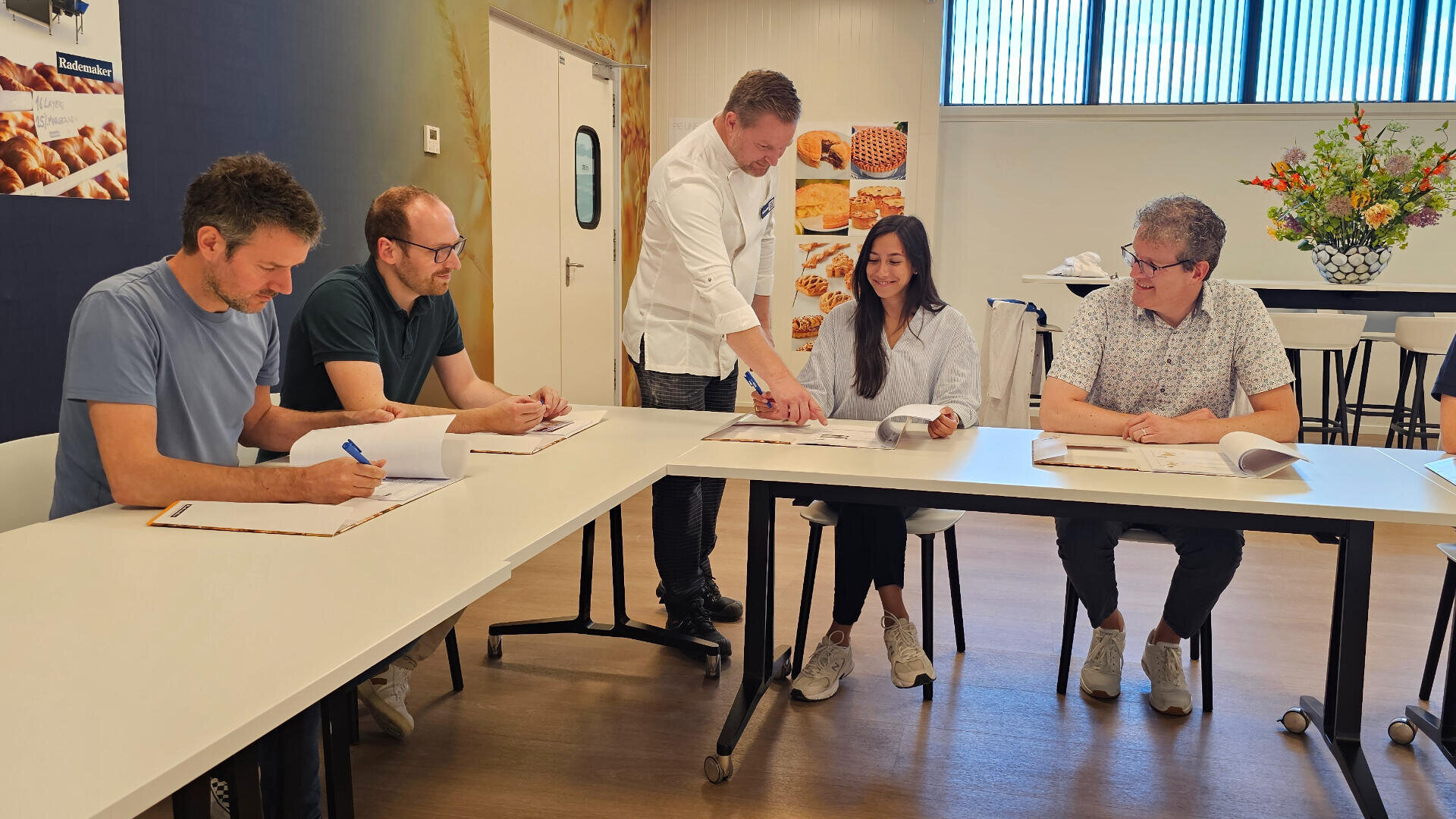 Five people sit around a table in a modern meeting room. A man standing points to a document while the others, seated, look on and take notes. There are baked goods images on the wall and a flower arrangement in the background.