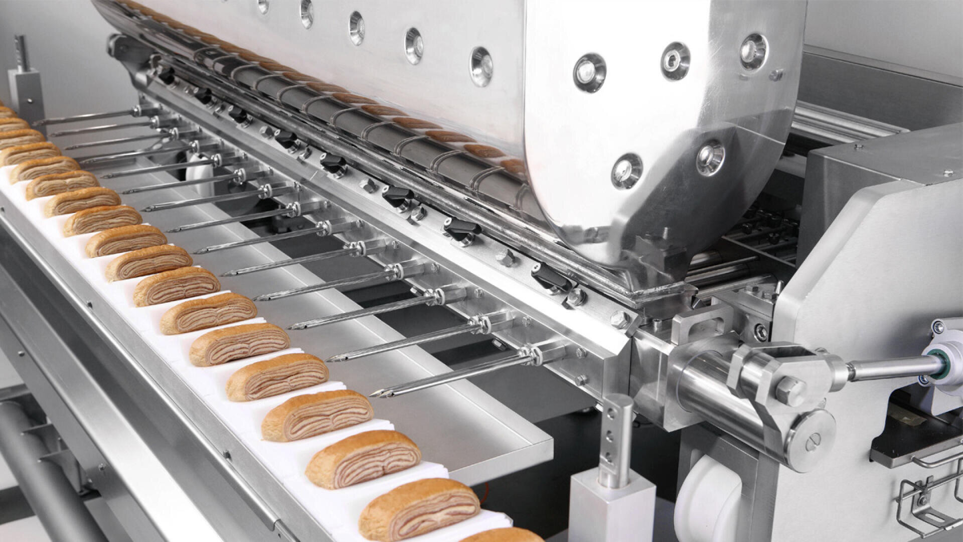 A close-up of an industrial bakery machine placing rows of layered pastries onto a conveyor belt in a food production facility.