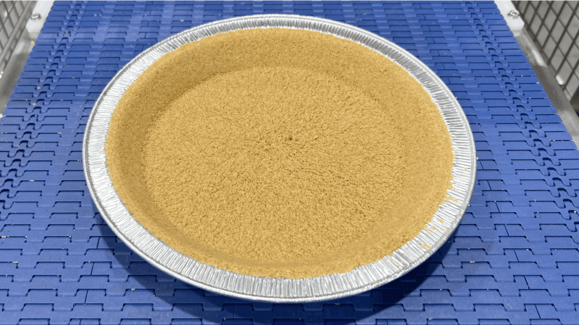 Ein einfacher Graham-Cracker-Kuchenteig in einer runden Aluminiumform liegt auf einem blauen Förderband, wahrscheinlich in einer Fabrik oder einer Großküche.