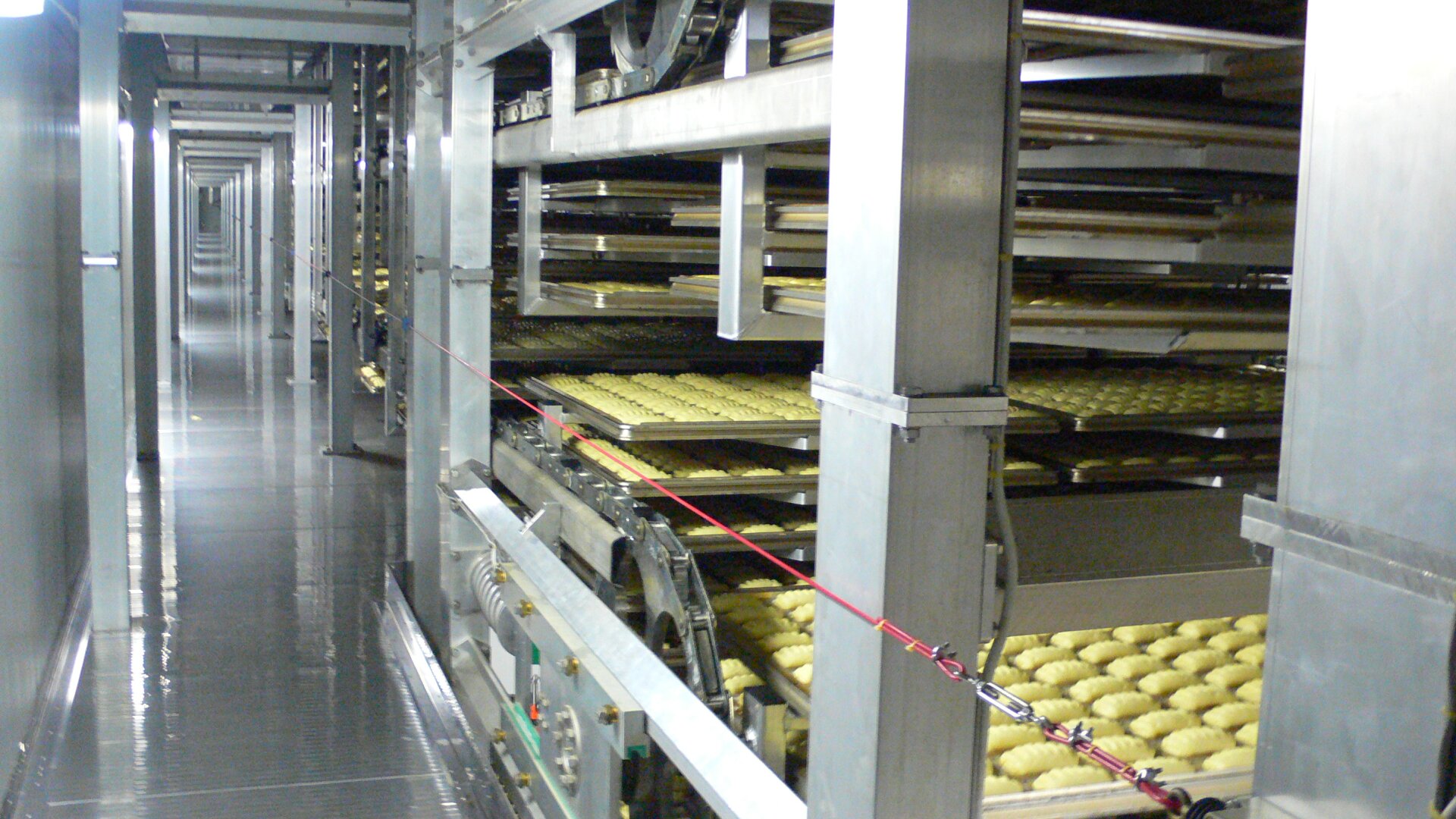 Rows of dough rise on trays in a large industrial bakery conveyor oven, with metal machinery and platforms surrounding the baking process.